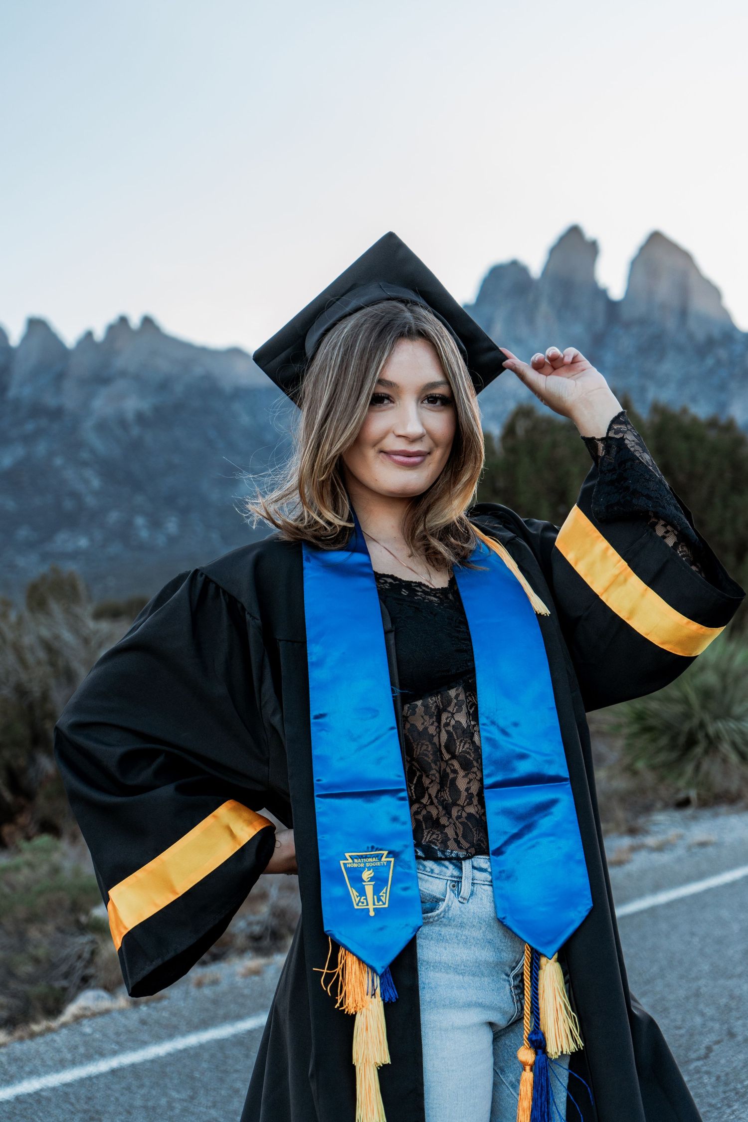 Graduate in black cap and gown touches tassel while standing on road with rocky mountains behind.
