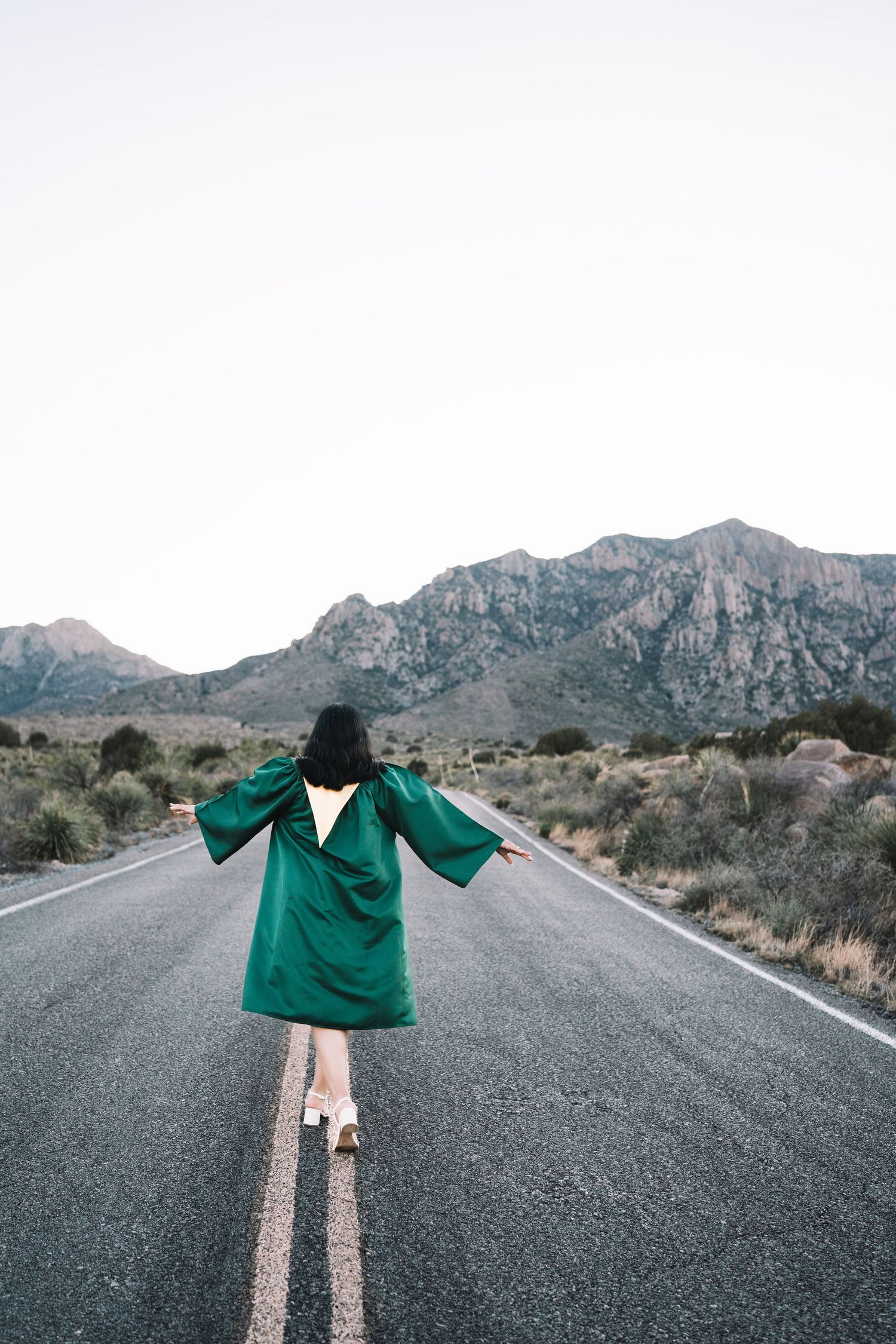 Female graduate in a green cap and gown walking with arms outstretched on an open desert highway.