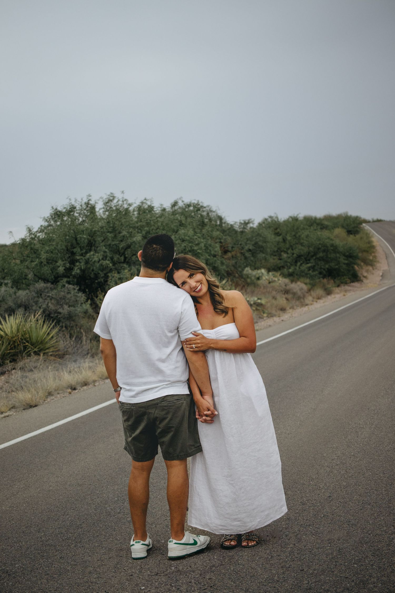 Woman in white dress leaning on mans shoulder smiling while holding hands on a desert road.