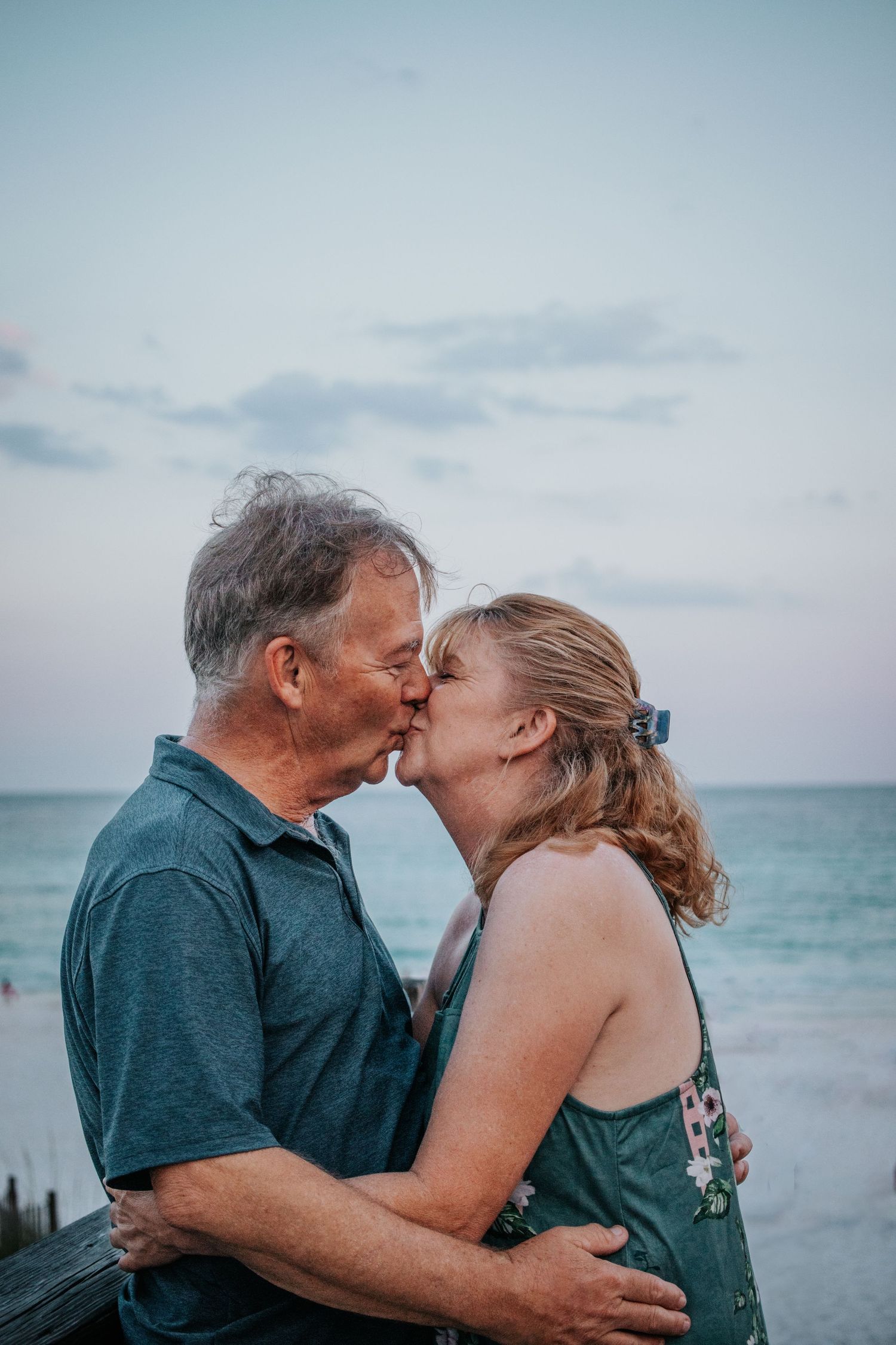 Older couple sharing a romantic kiss on the beach at sunset with turquoise ocean water in the background.