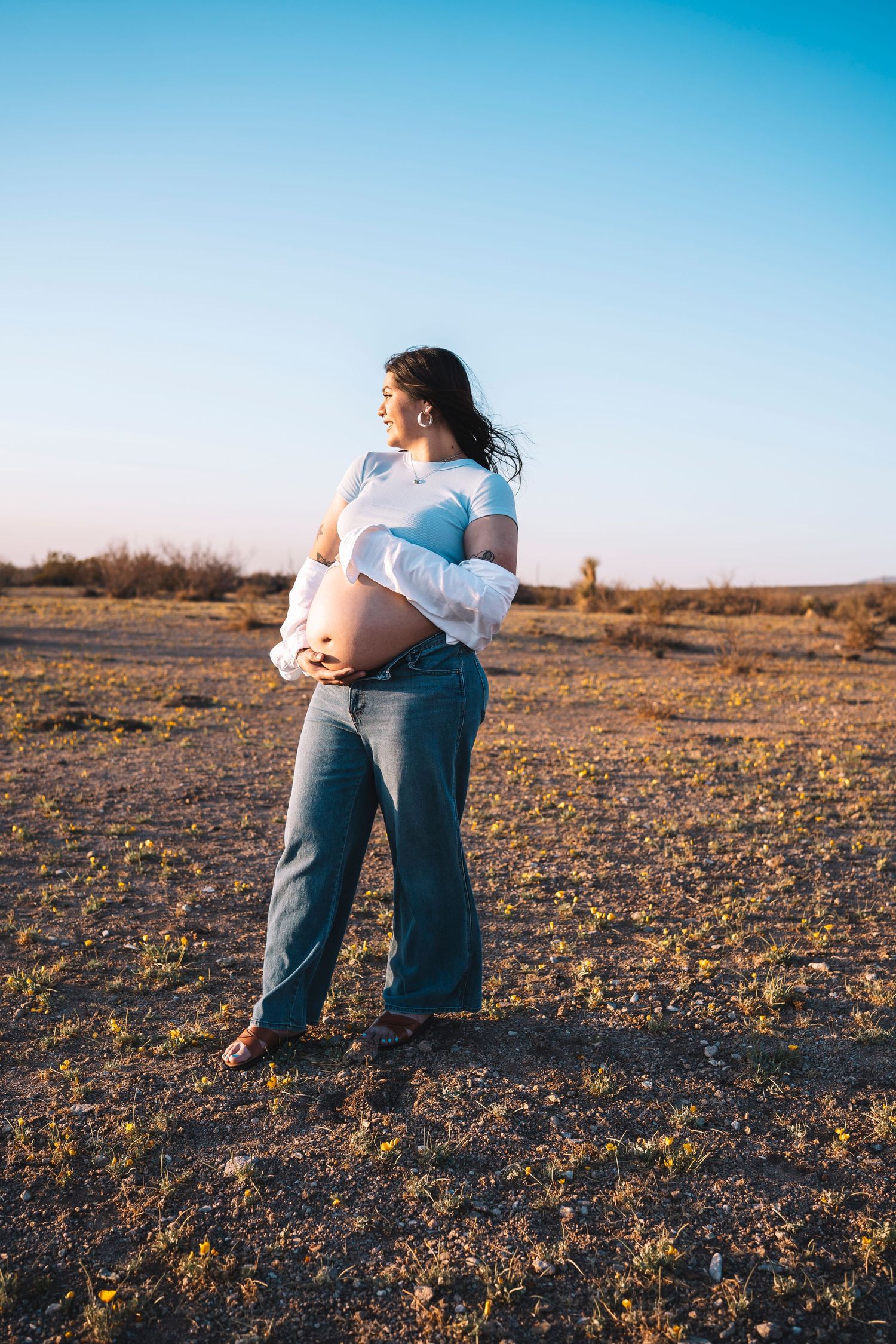 Pregnant woman in blue crop top, white shirt, and jeans cradling her baby bump outdoors in a desert landscape at sunset.