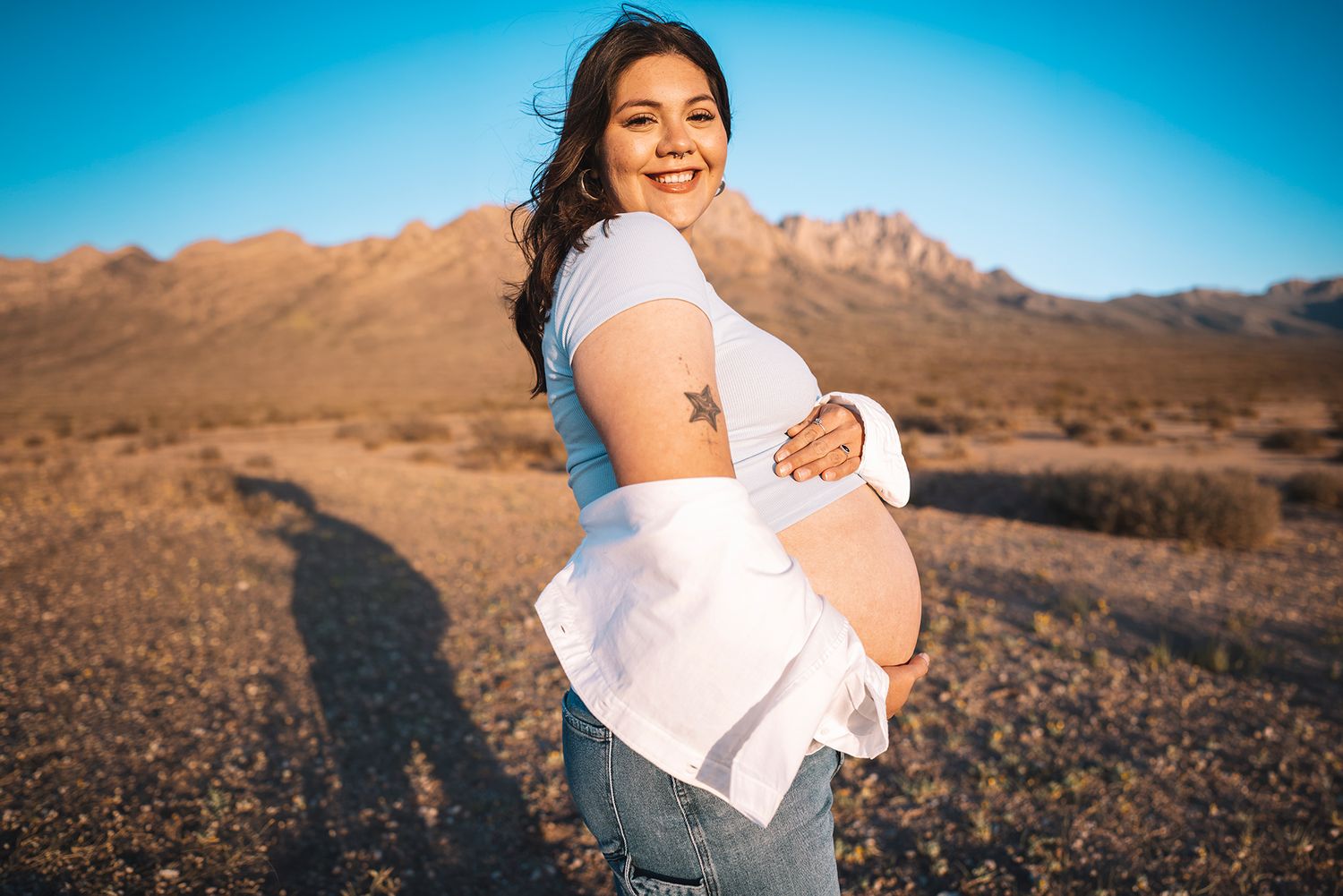Cheerful pregnant woman in white shirt and jeans shows baby bump in sunny desert setting.