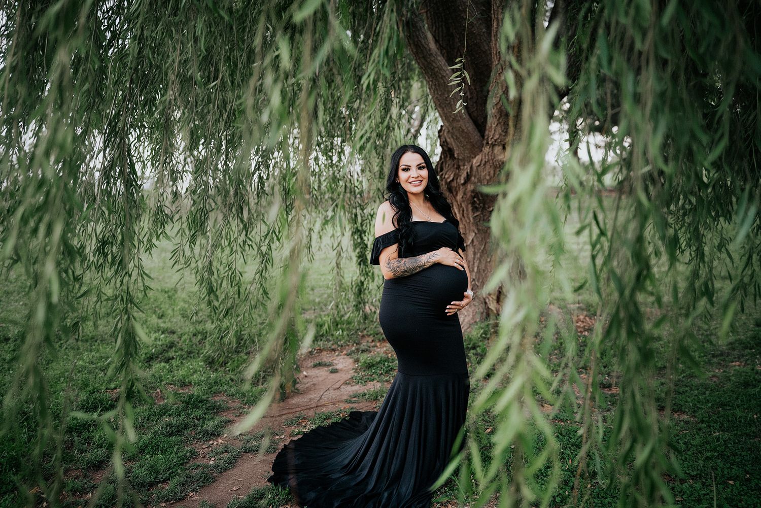 Smiling pregnant woman in black dress posing under weeping willow tree outdoors.