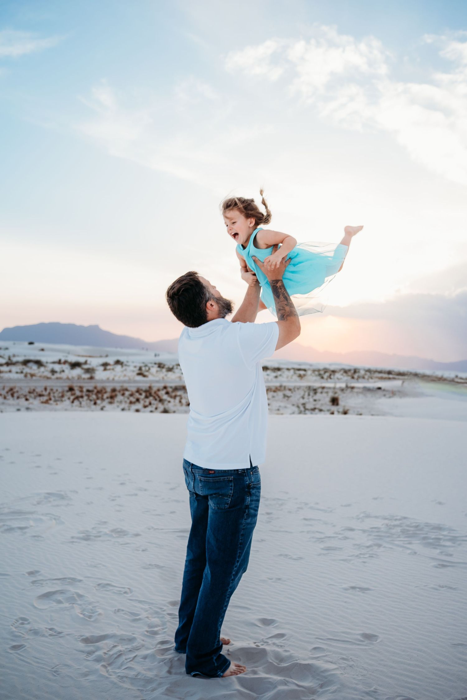 Father lifting young daughter in turquoise dress high in the air on white sand dunes at sunset.