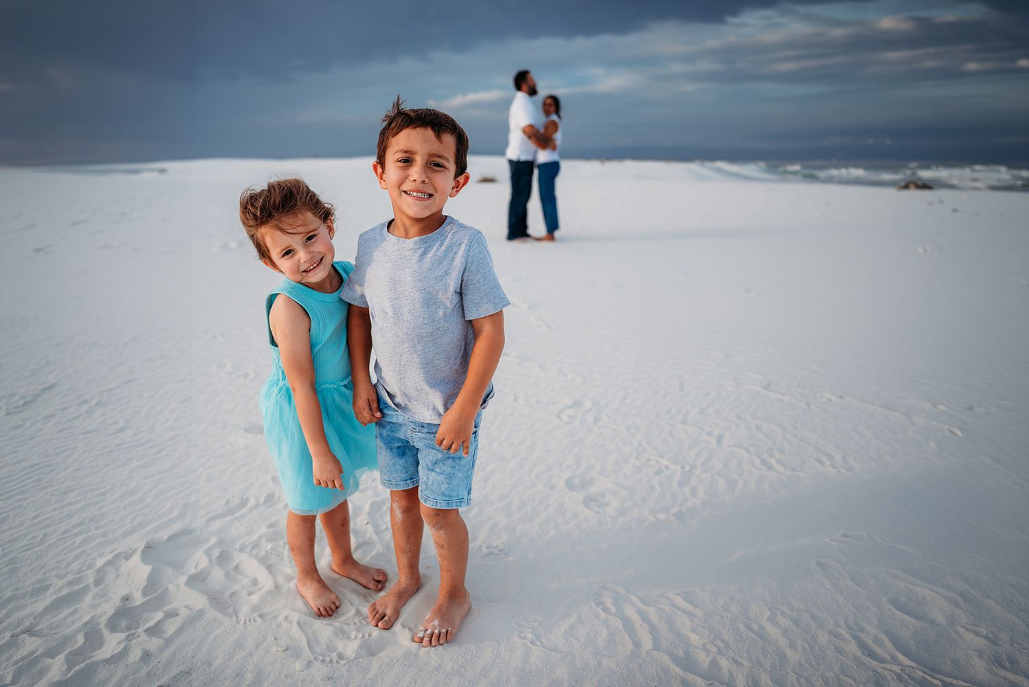 Two young children smile and pose on white sand dunes with their parents standing in the background.