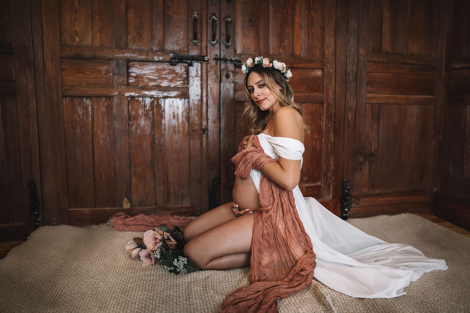 Pregnant woman in white dress and floral crown sits gracefully on floor before rustic wooden doors.