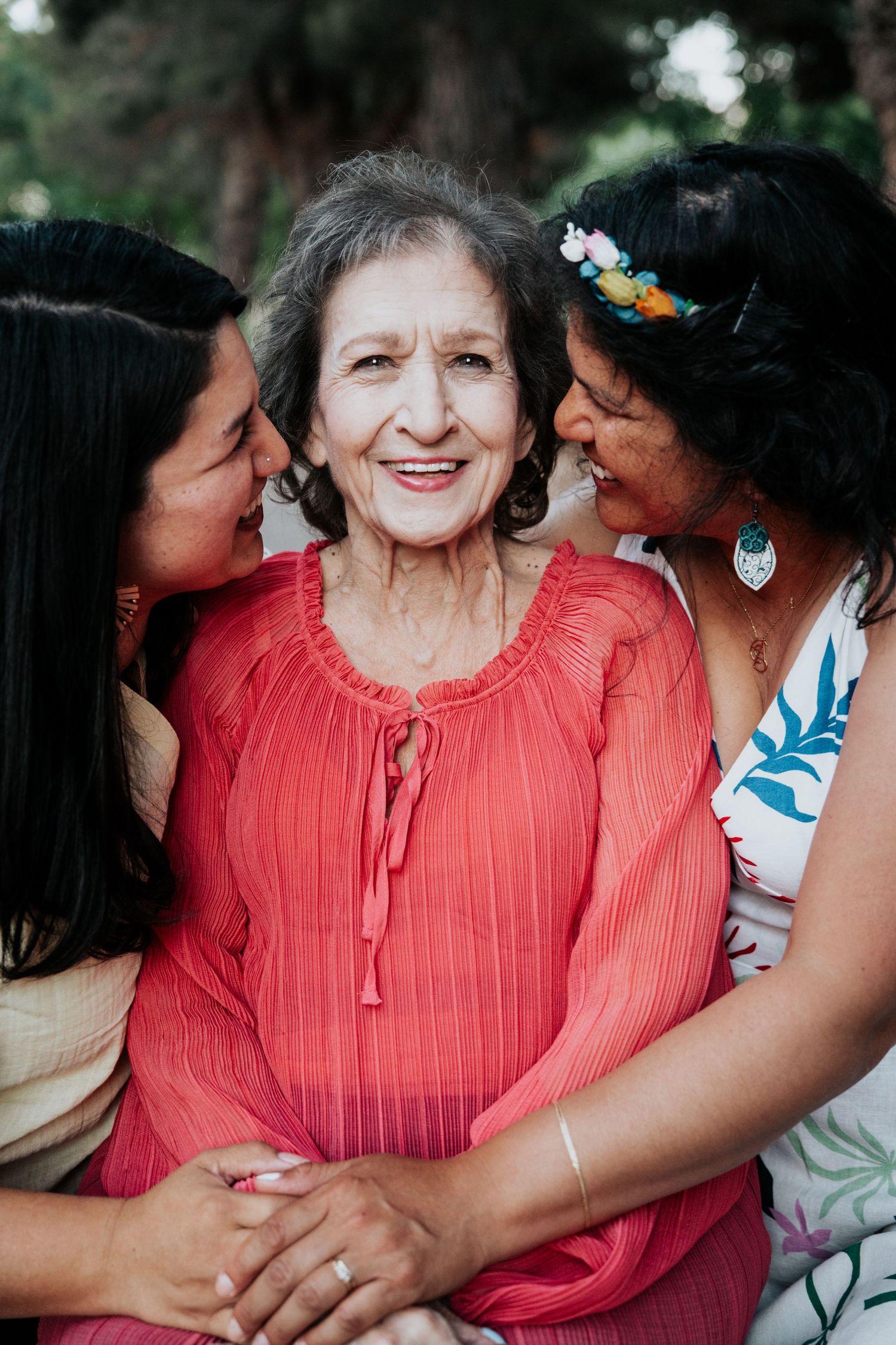 Two younger women lovingly kiss the cheeks of a smiling elderly woman in a red blouse outdoors.