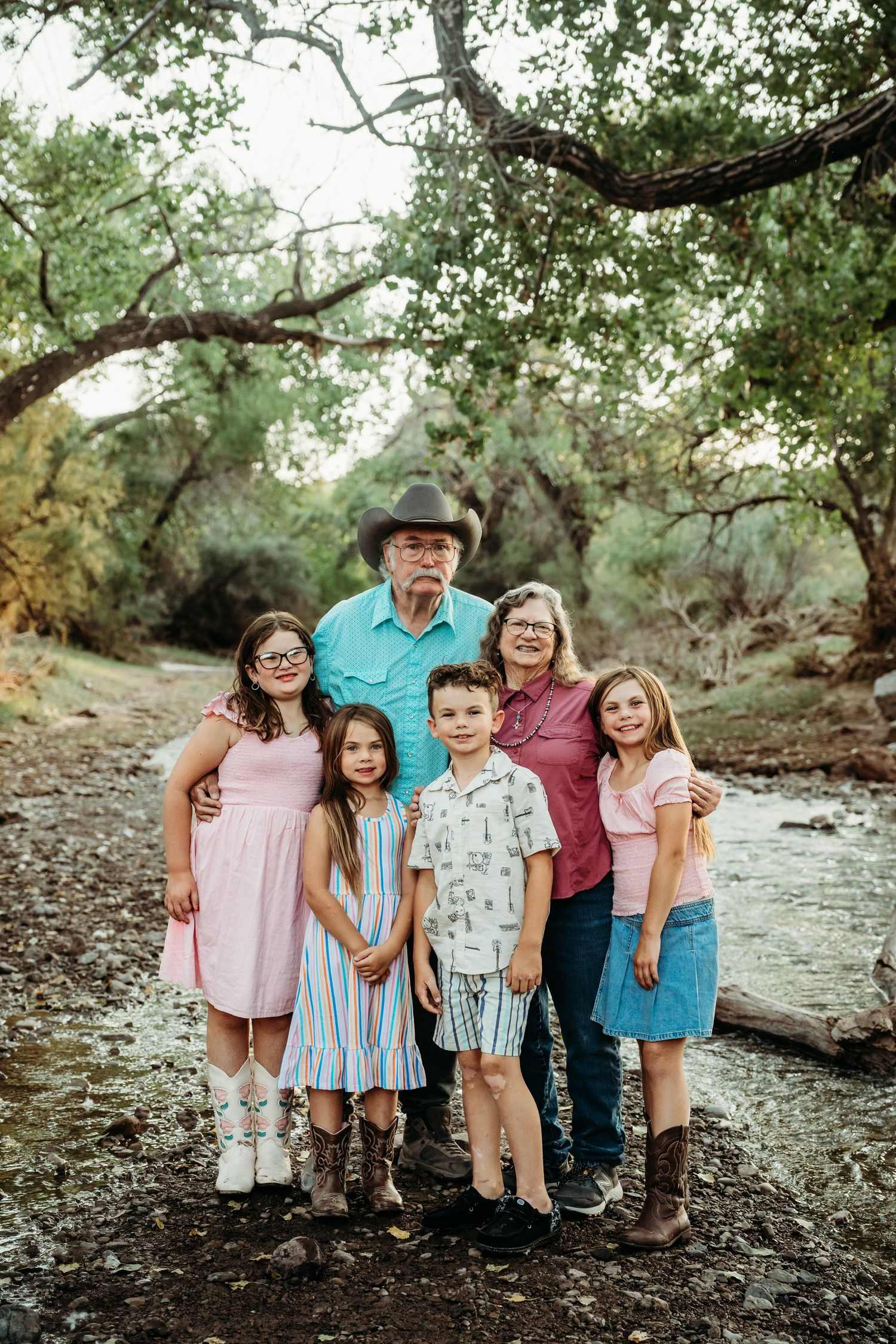 Grandparents in western wear pose with four grandchildren near a tree-lined creek for a family portrait.