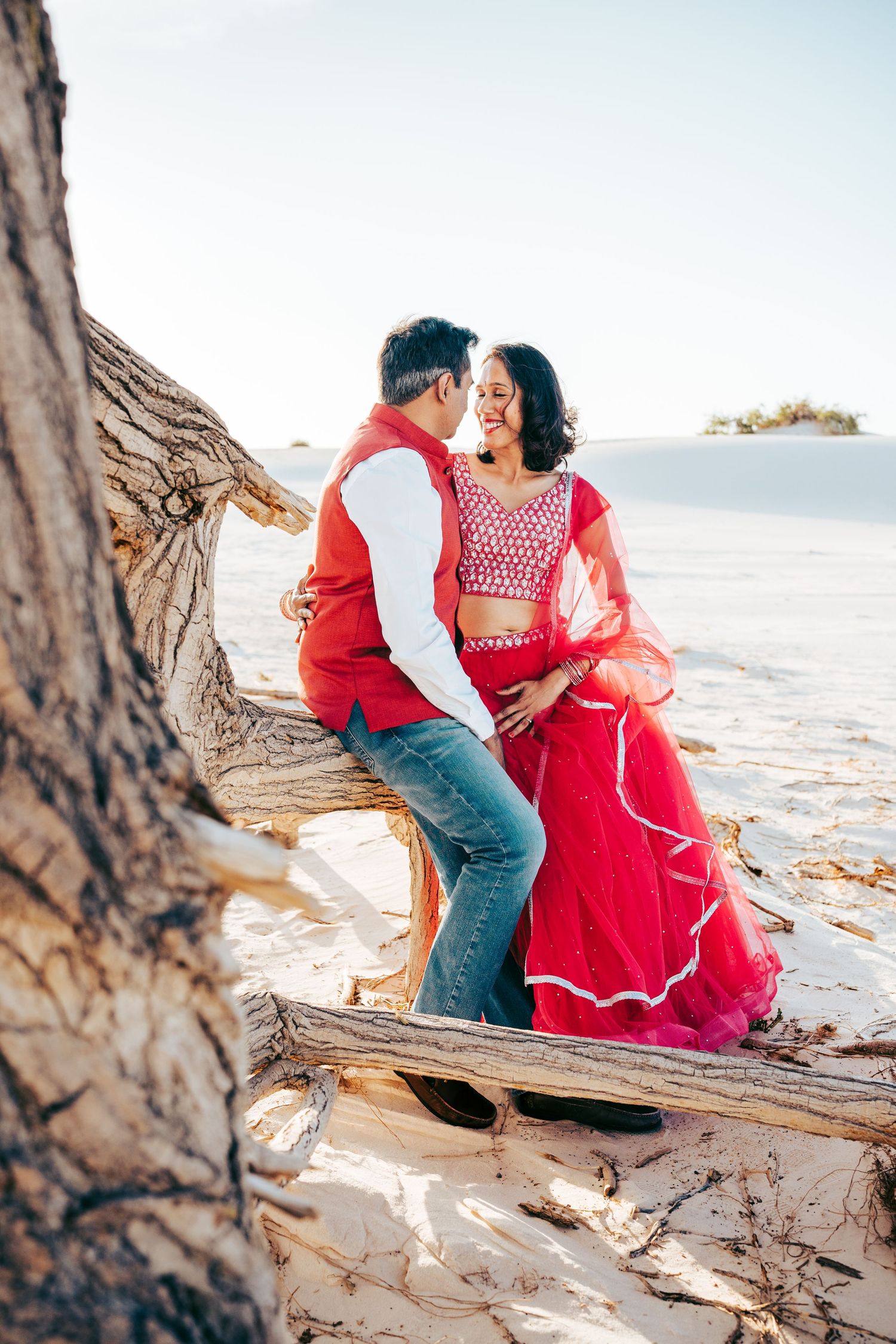 Indian couple in traditional red and white clothing sharing an intimate moment on driftwood.