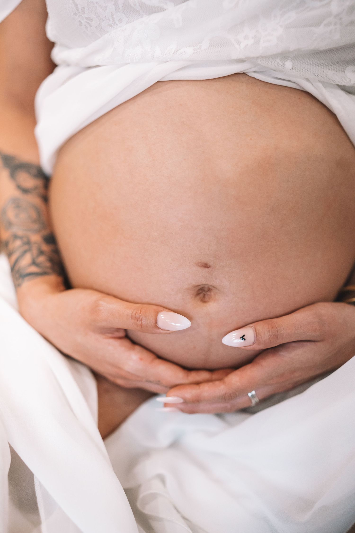 Close-up of pregnant belly cradled by hands with white manicure, tattoo visible on side.