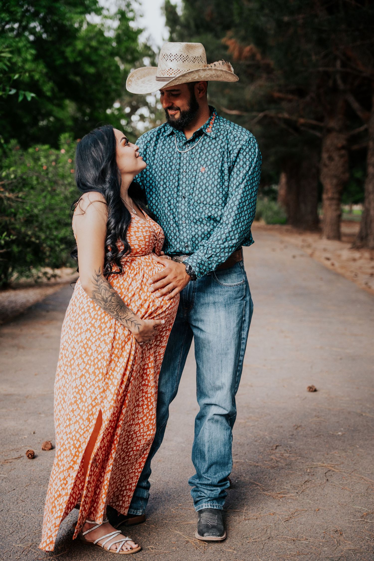 Smiling pregnant woman in orange floral dress leans into partners embrace, man wearing cowboy hat and teal shirt.