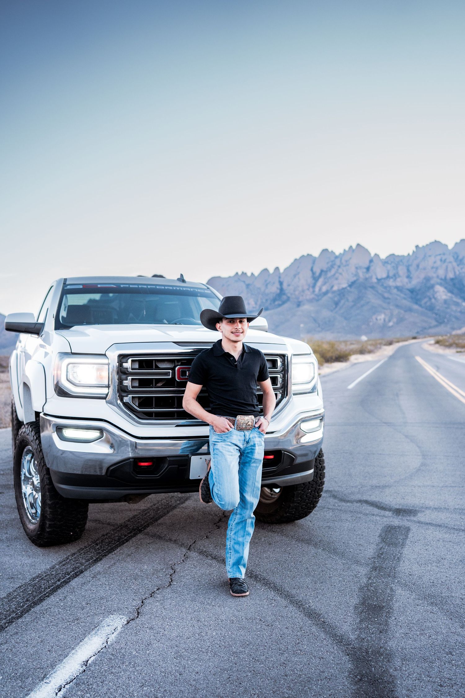 Cowboy in black hat leans against white GMC pickup truck on desert highway with mountain backdrop.