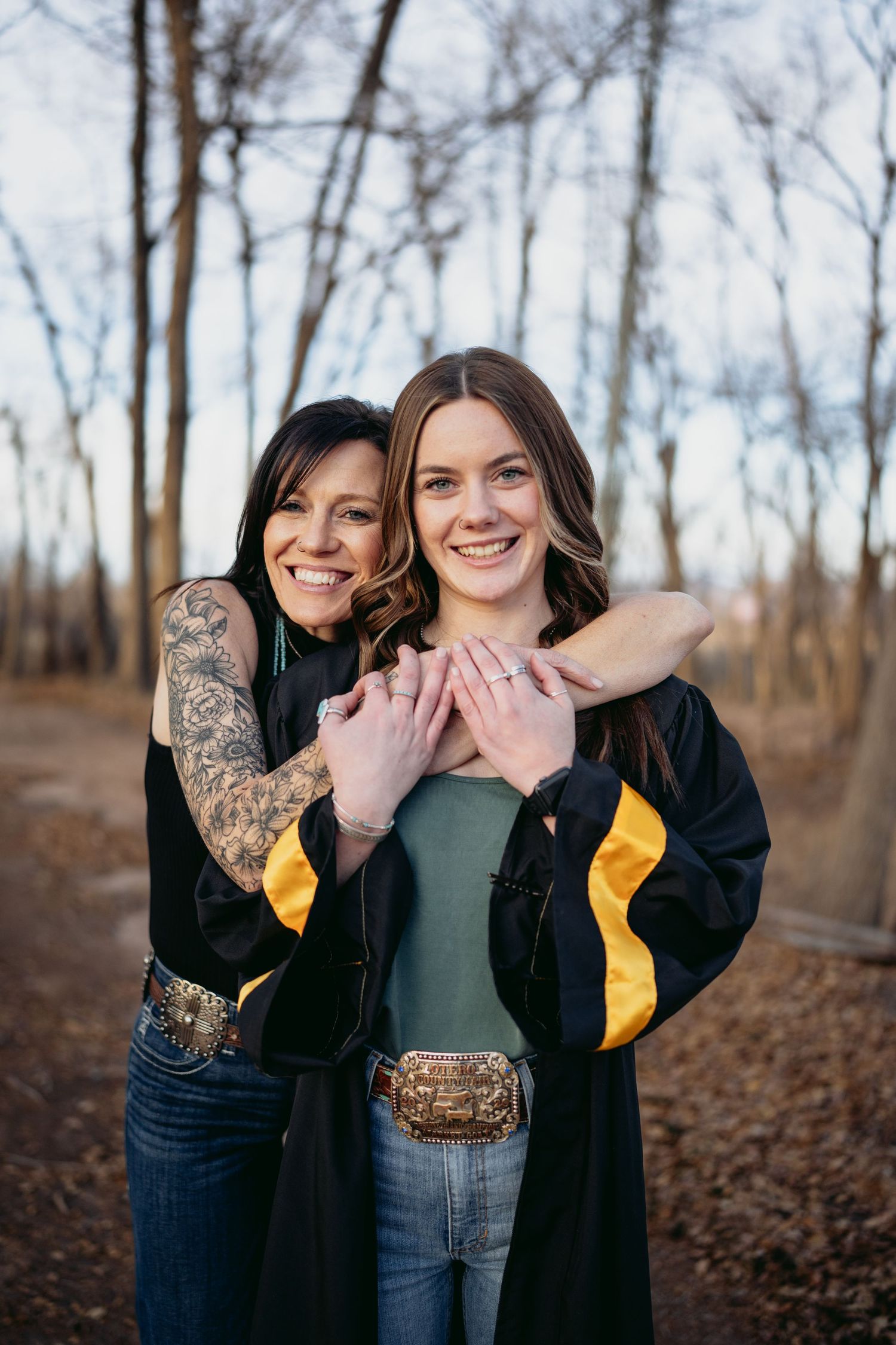 Two smiling women in black graduation gown embracing outdoors among bare winter trees.