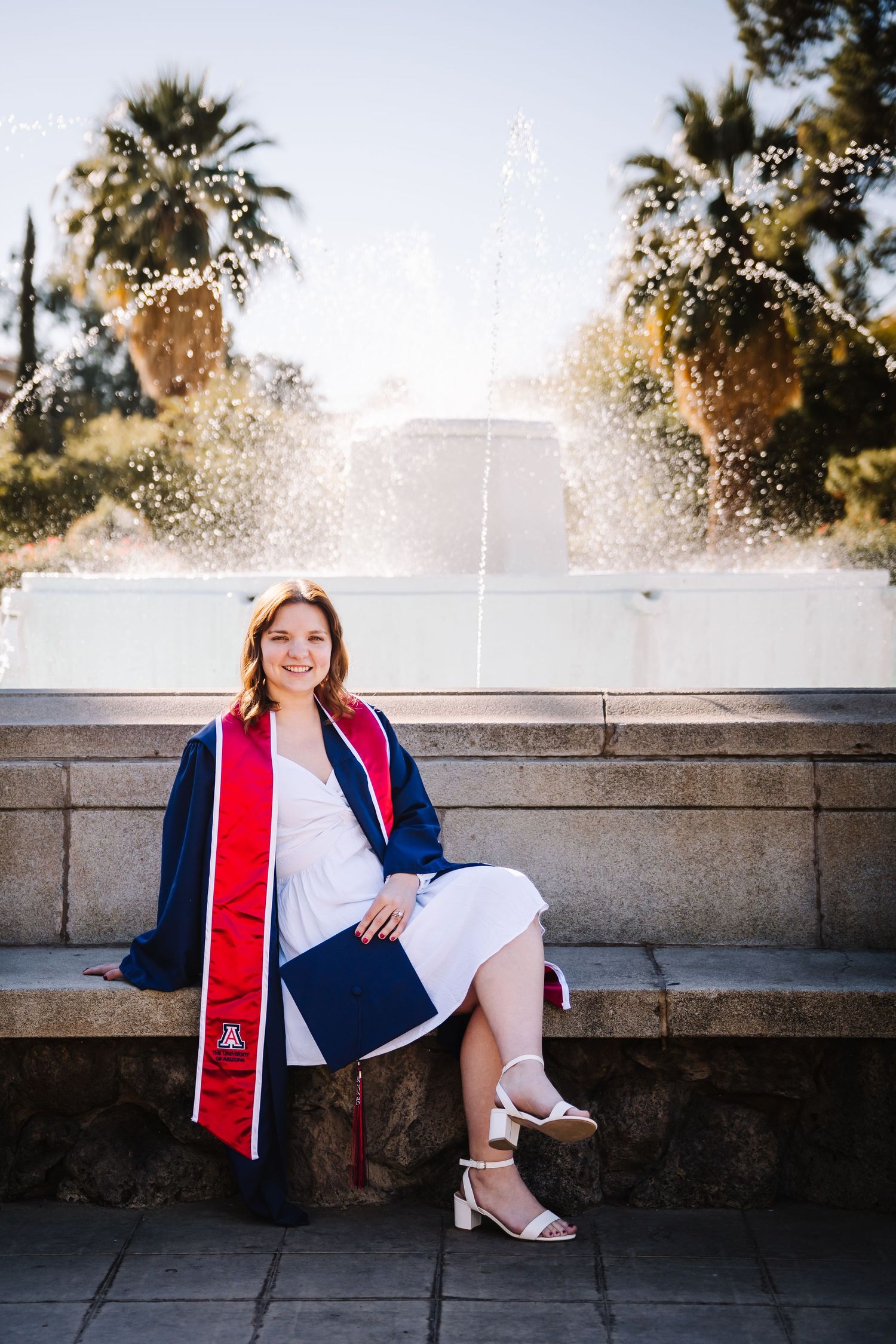 Smiling graduate in navy cap and gown with red stole sits by fountain holding diploma on sunny day.