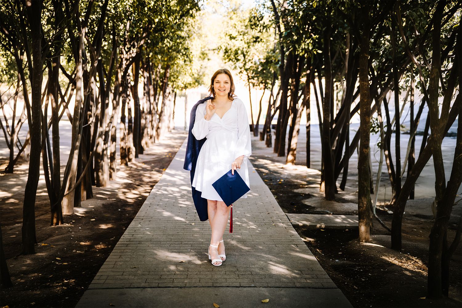 Woman in white dress walks on a tree-lined boardwalk holding a navy clutch on a sunny day.