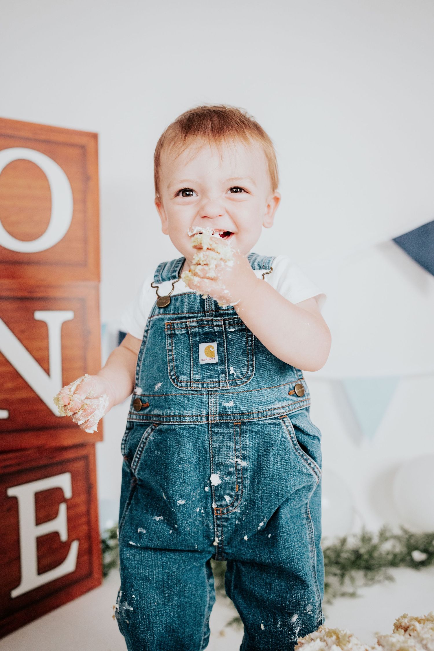 Adorable baby in denim Carhartt overalls eating cake during first birthday smash cake photo session.