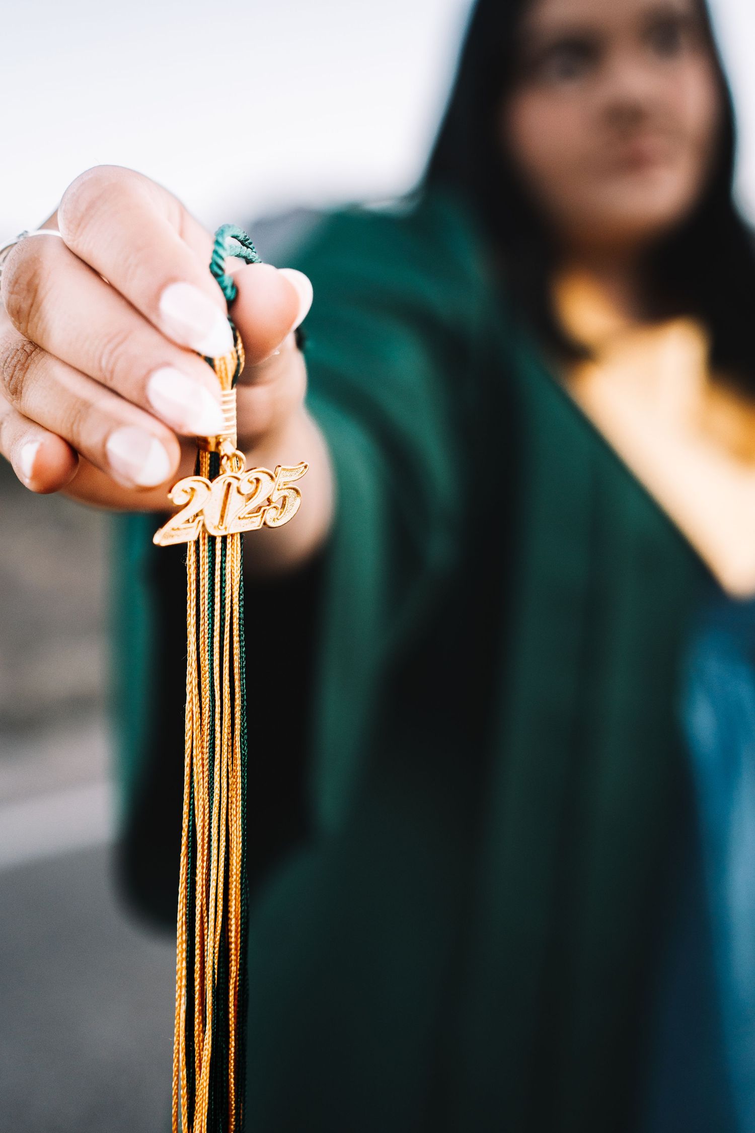 Close-up of hand holding gold 2025 graduation tassel toward camera with blurred graduate in background.