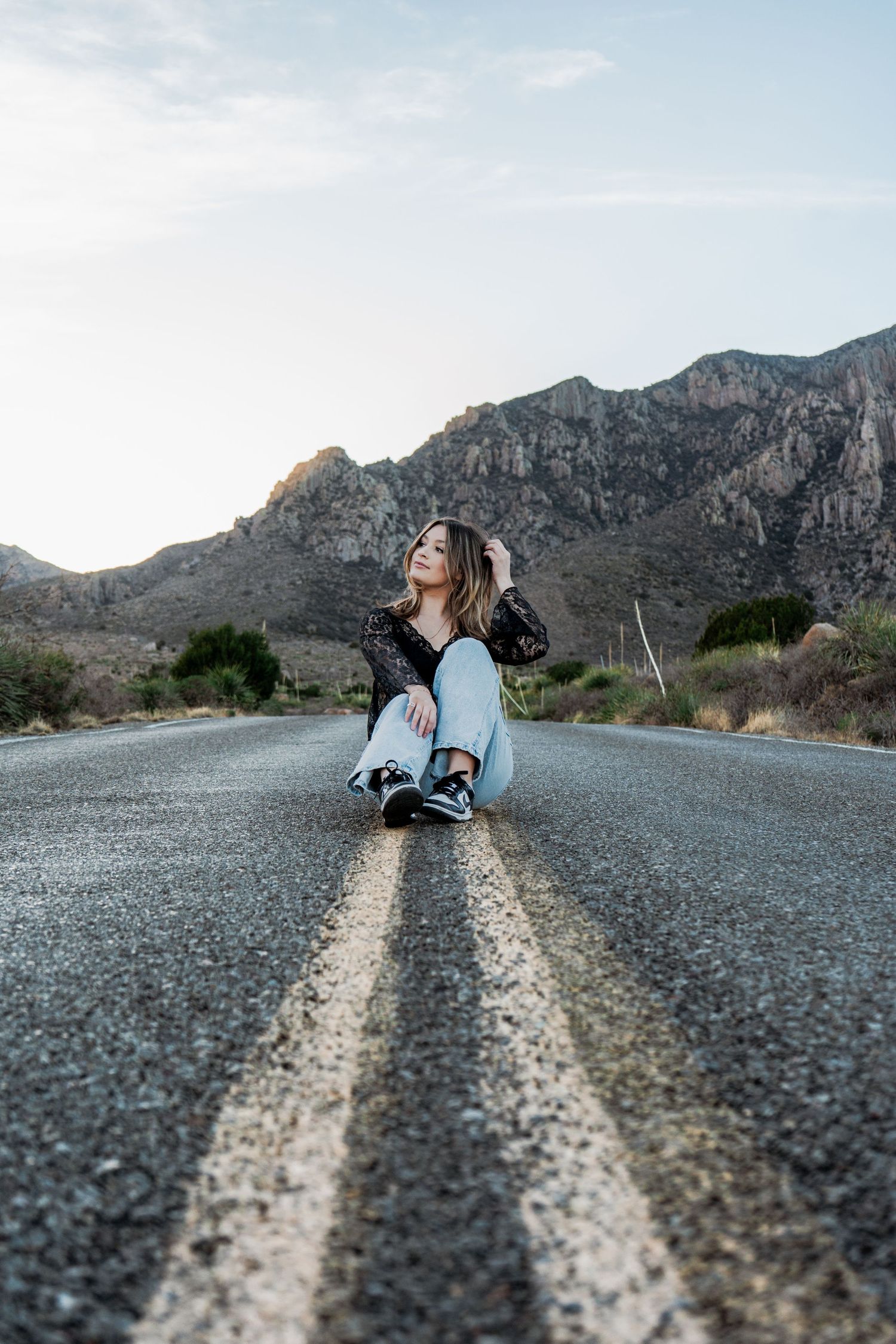 Young woman in black lace top sits on road centerline gazing upward with mountains behind.