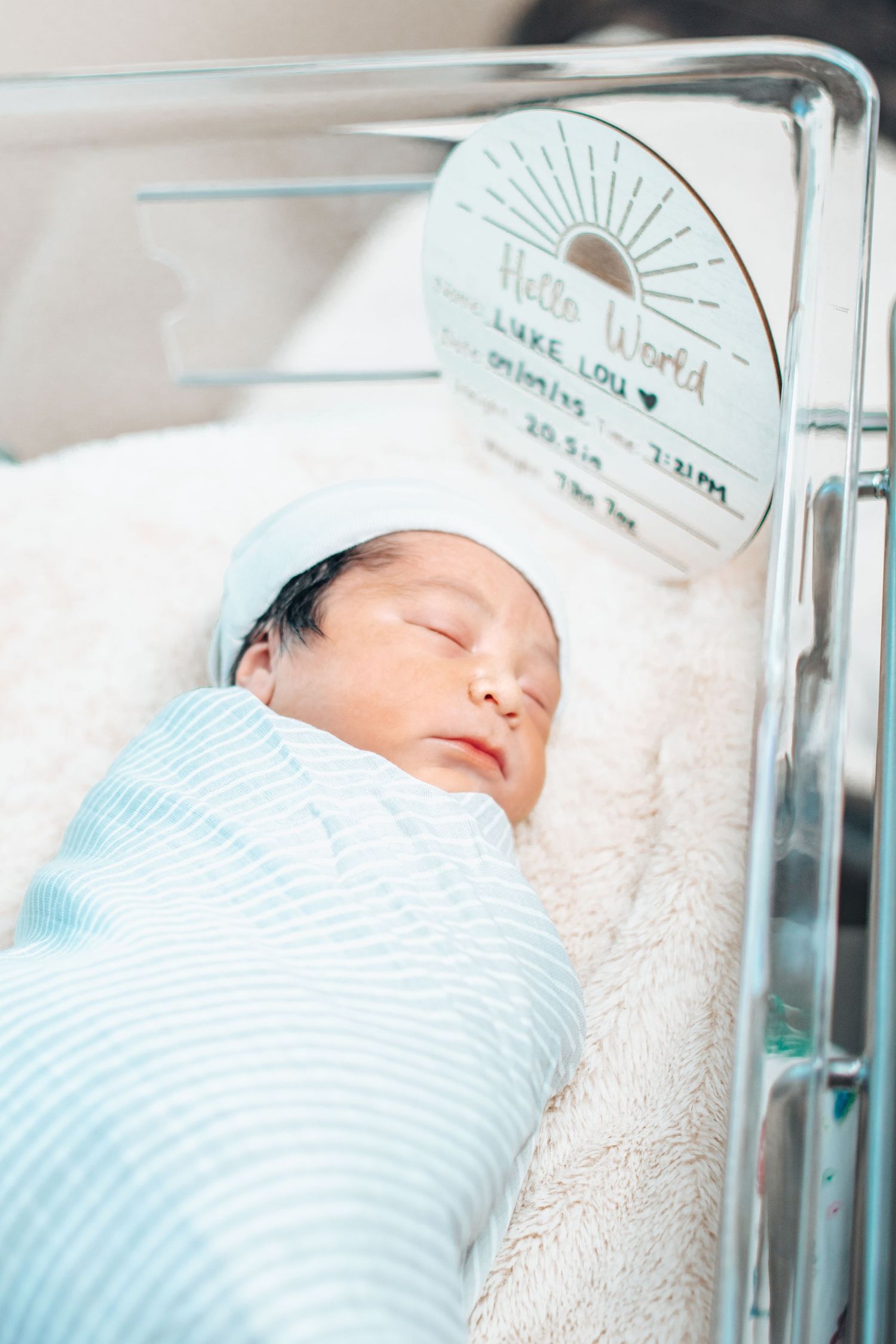 Sleeping newborn baby Luke Lou swaddled in blue striped blanket in hospital bassinet with birth card.