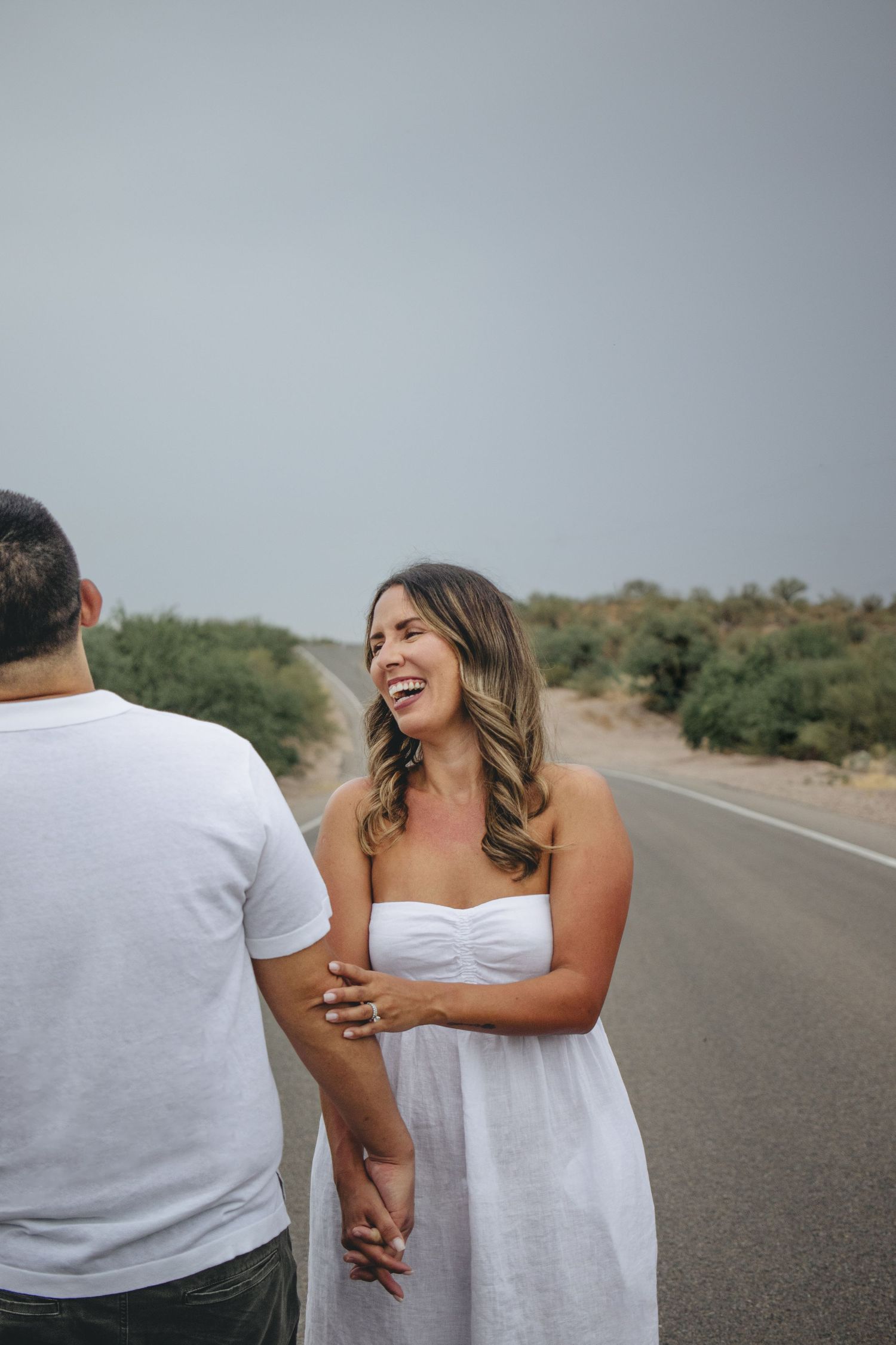 Happy woman in white dress laughing while holding hands with man on empty desert road.
