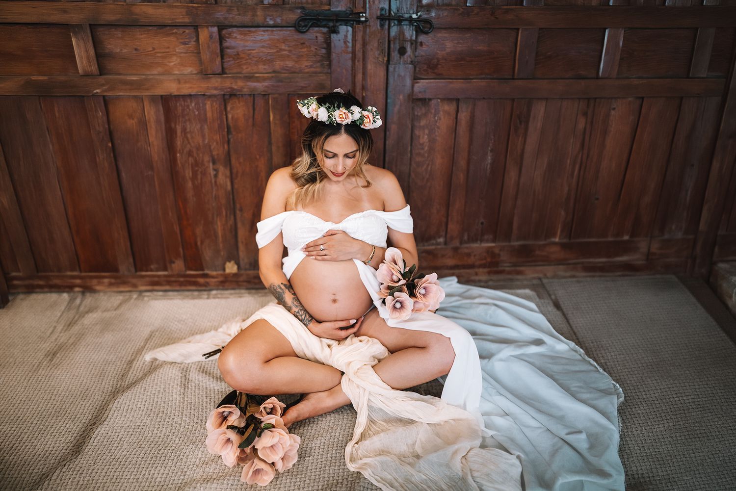 Pregnant woman wearing floral crown and white outfit sits on floor holding flowers near wooden doors.