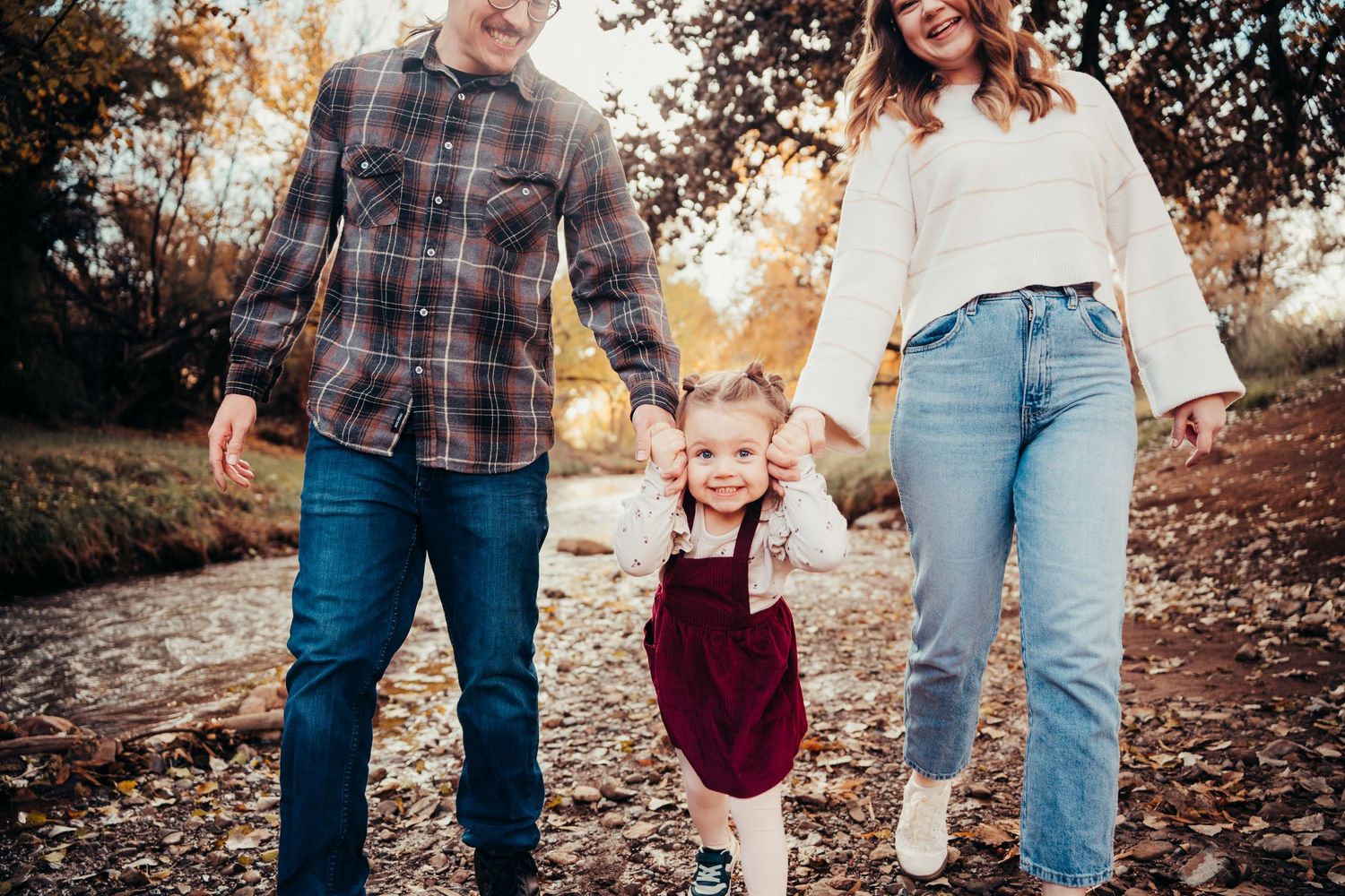Parents swinging their toddler daughter in a burgundy dress between them on an autumn creek path.