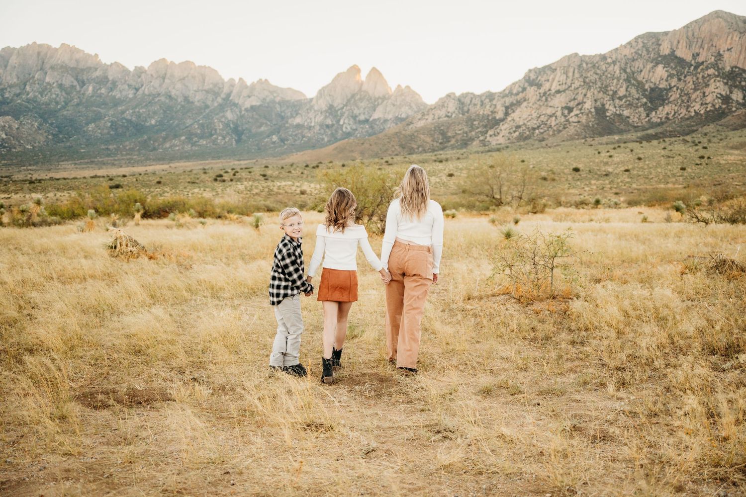 Mother and two children walk hand-in-hand through golden desert field toward scenic mountains.