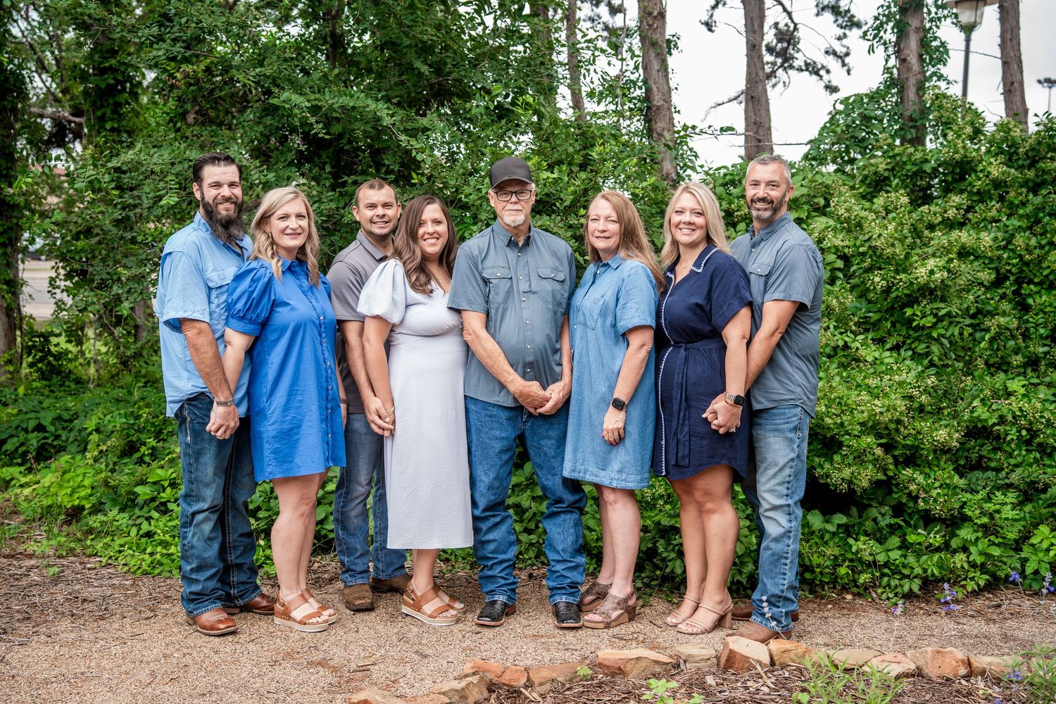 Extended family of eight adults pose together smiling outdoors among green trees and natural surroundings.