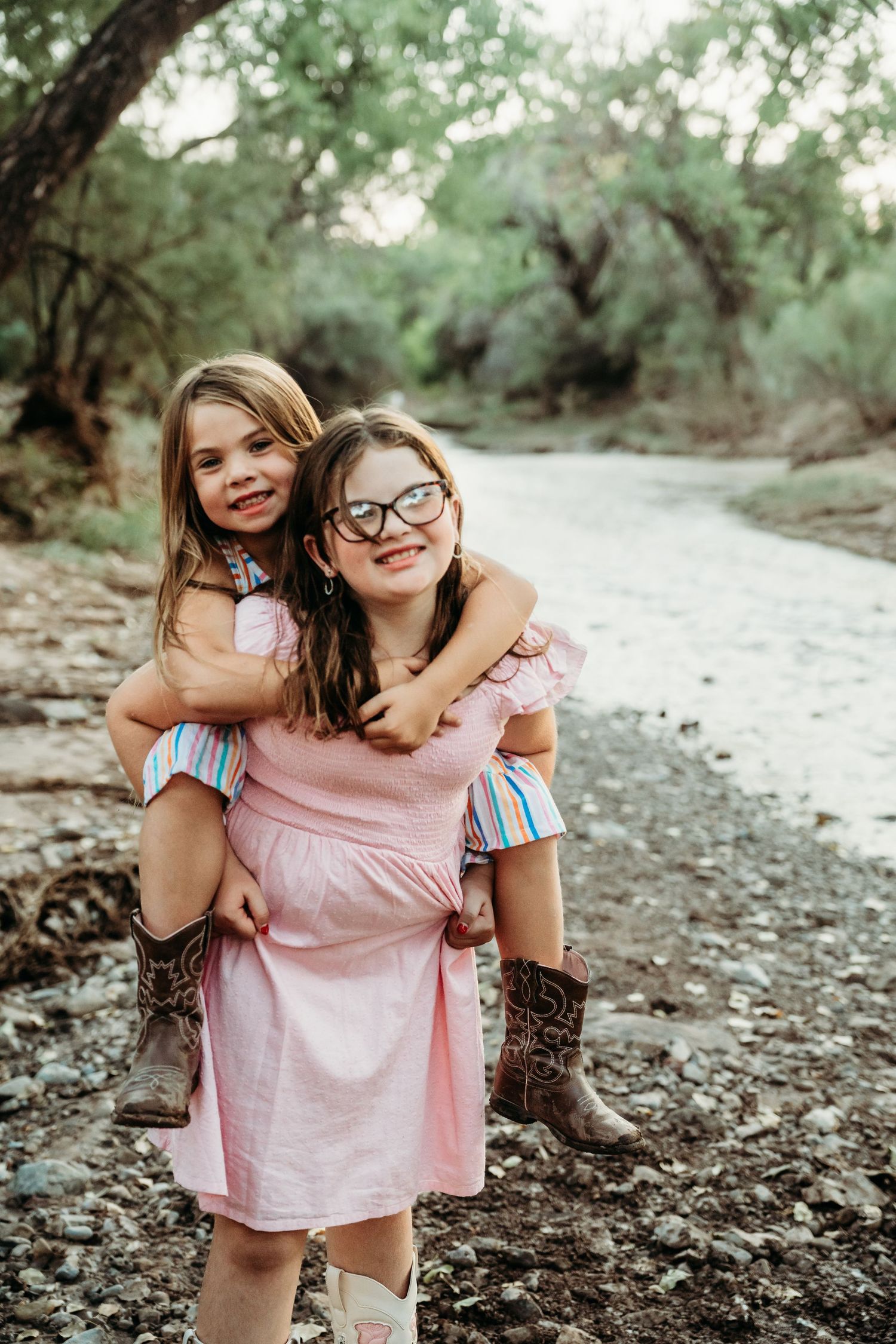 A girl in a pink dress giving her younger sister a piggyback ride along a rocky riverside.