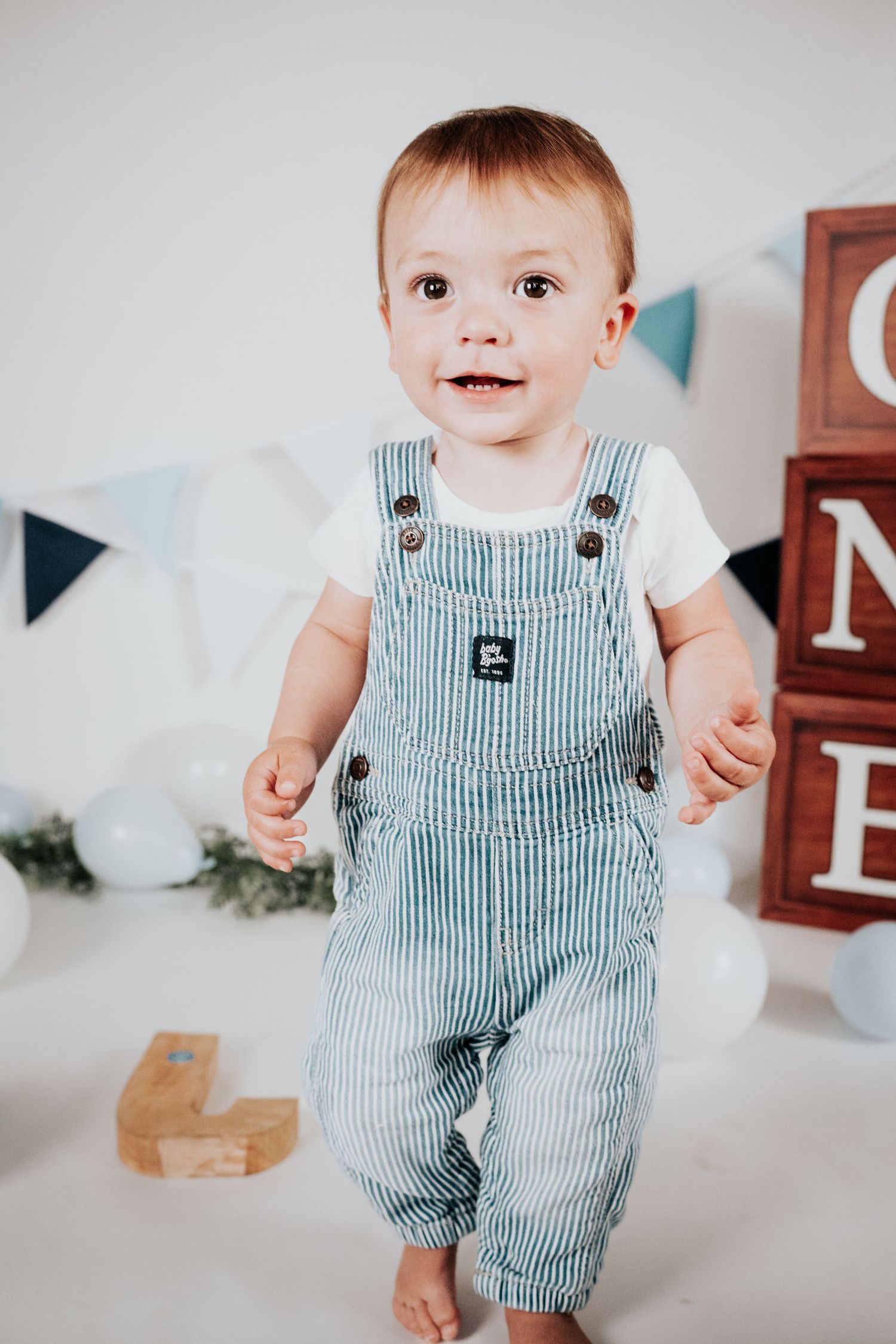 Adorable toddler in striped overalls standing at first birthday photoshoot with ONE blocks.
