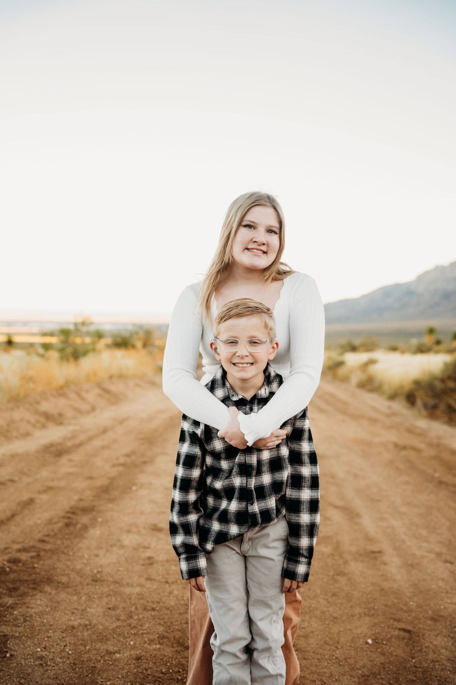 A smiling teenage girl hugs a young boy from behind on a desert dirt road.