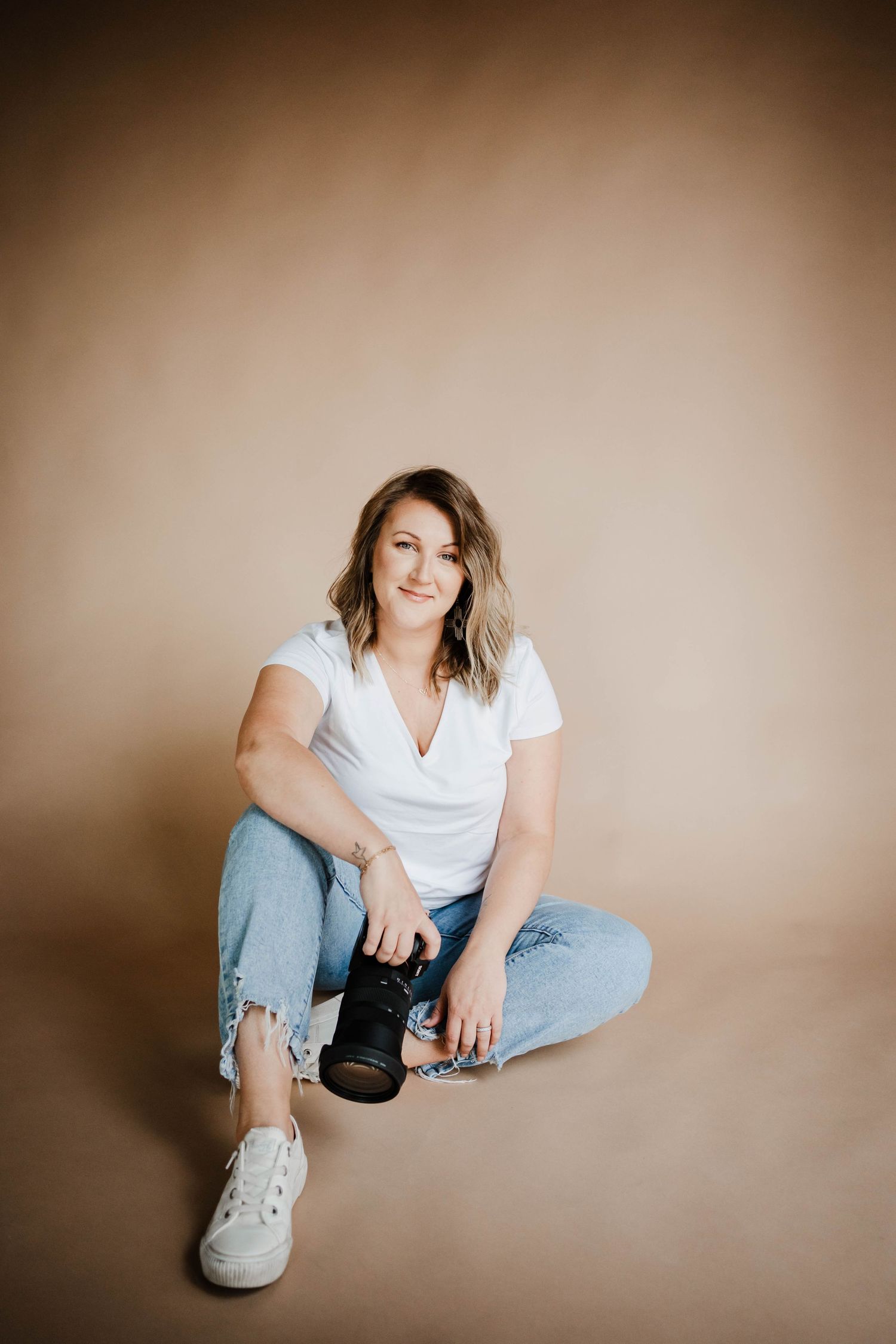 Female photographer sitting on floor holding camera, wearing white top and jeans on beige background.