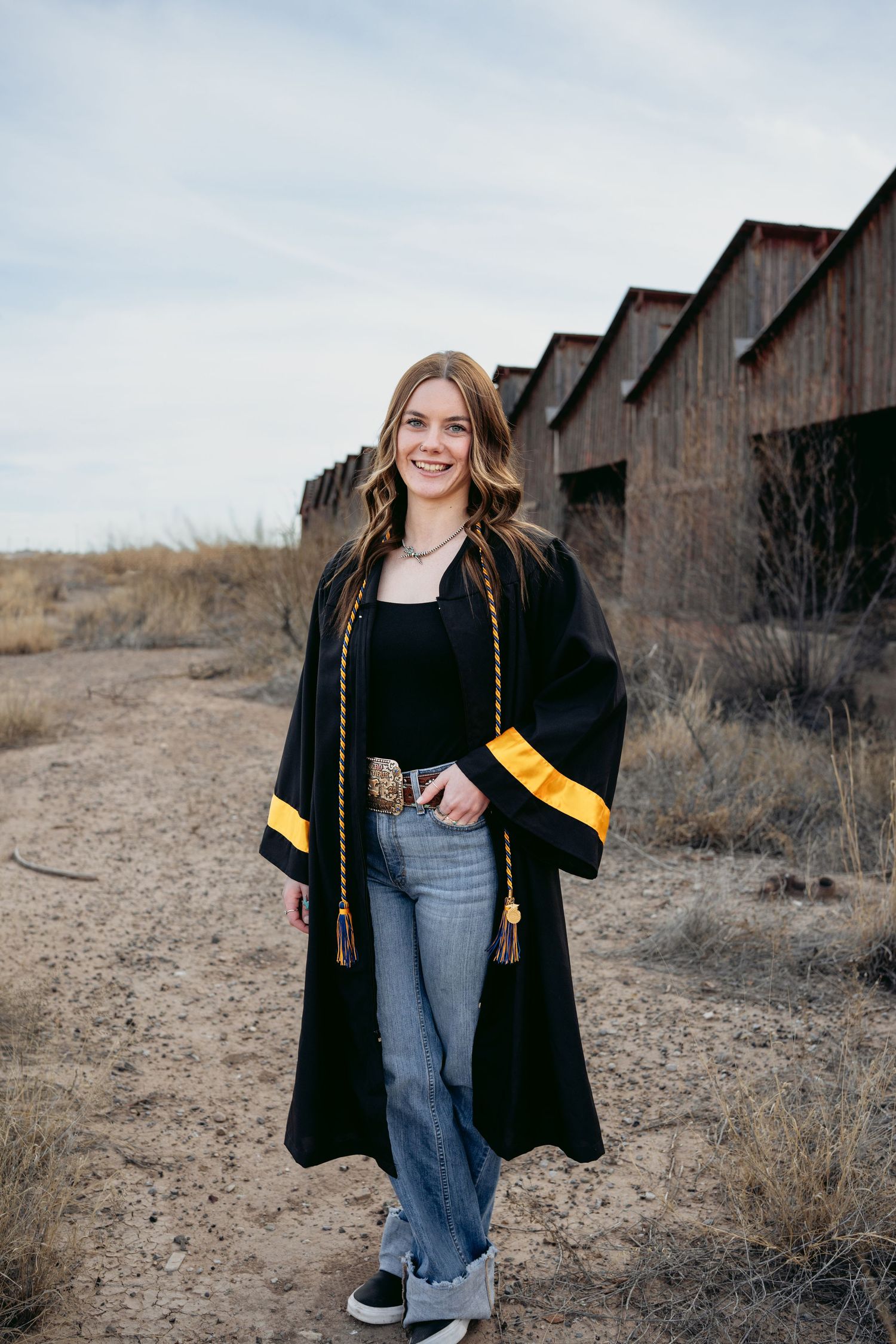 Smiling senior girl in black graduation gown with gold trim and braided cords outdoors near rustic barn.