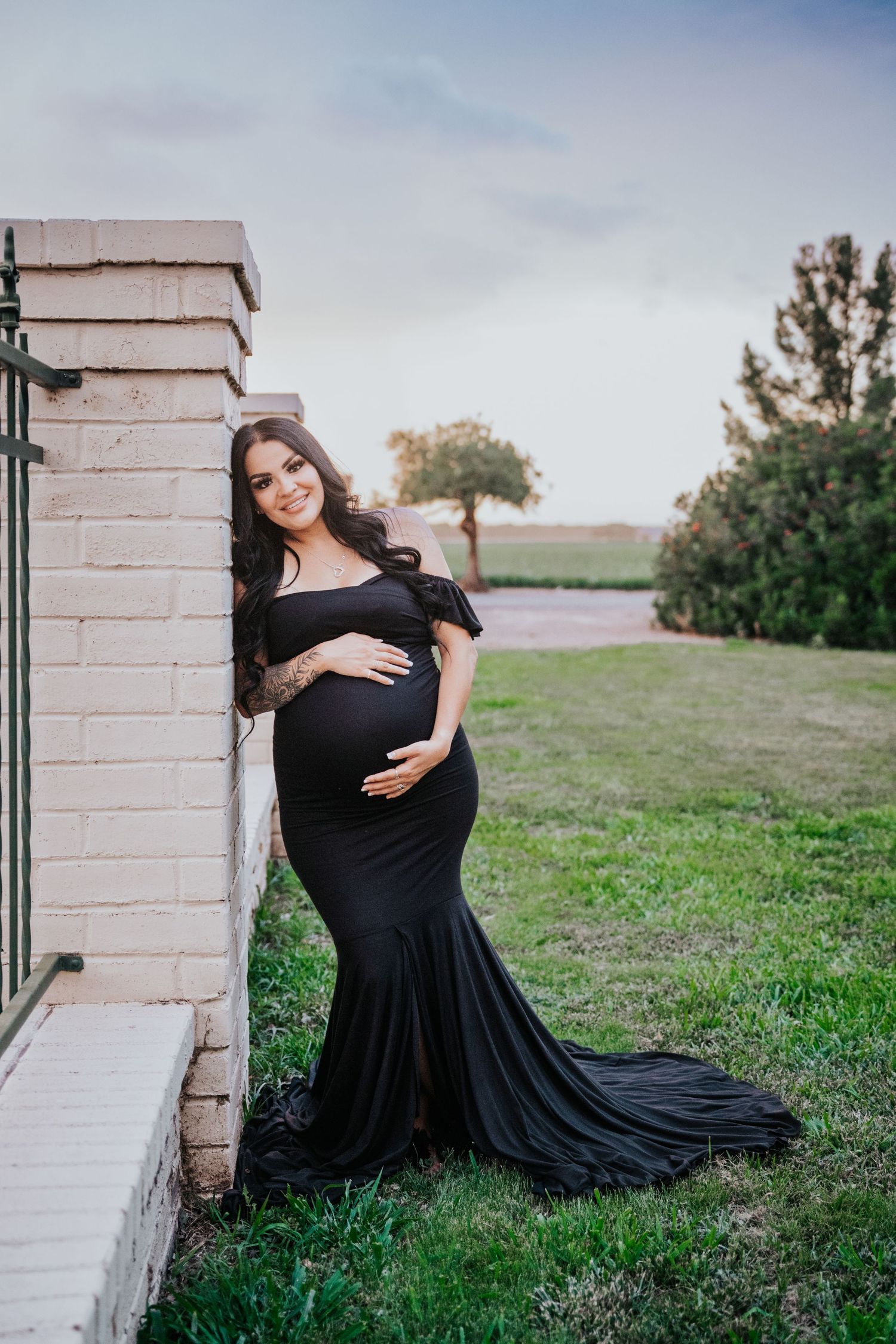 Pregnant woman in elegant black off-shoulder gown posing by brick pillar at sunset.
