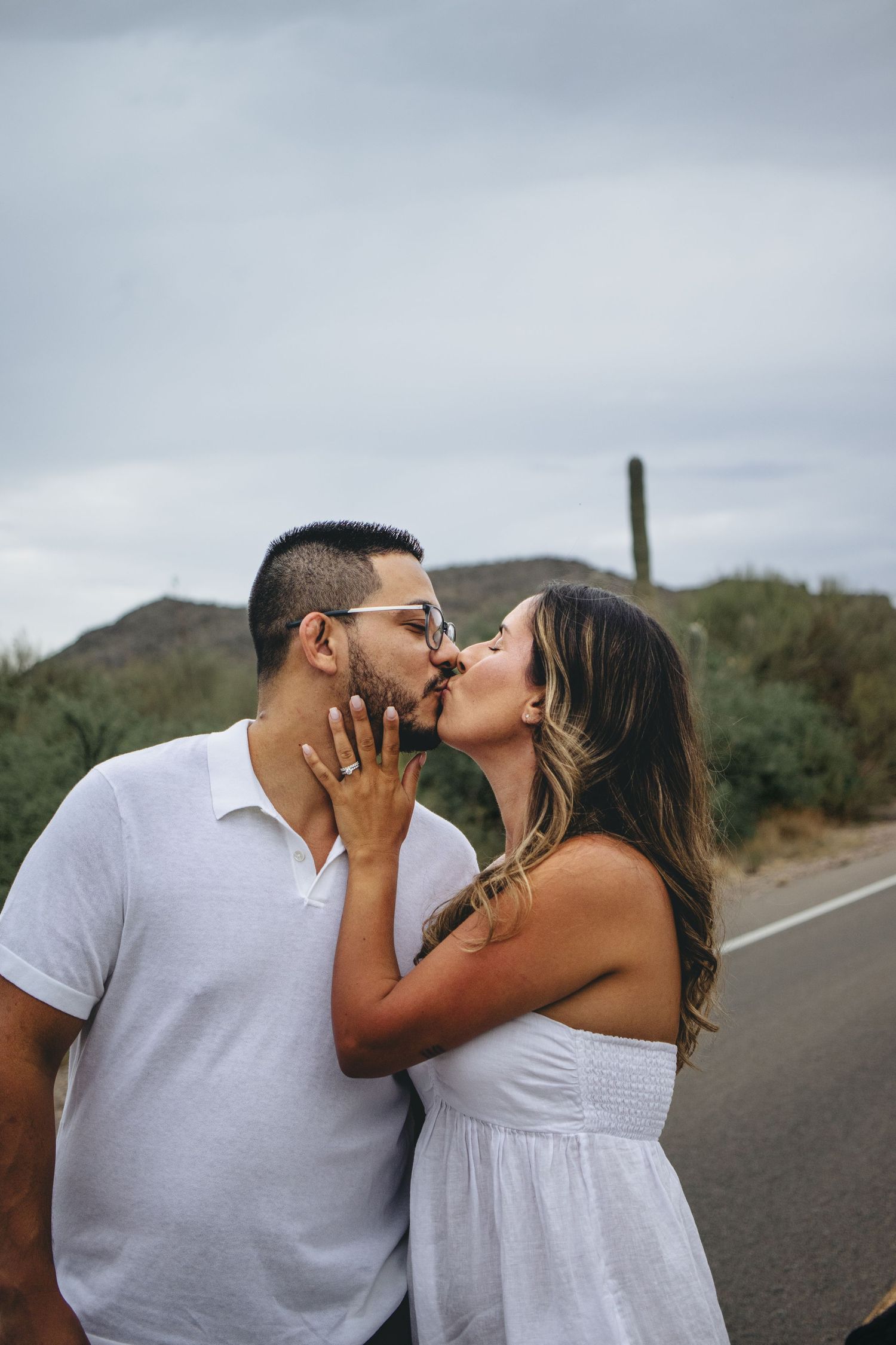 Couple sharing a kiss on a desert road with saguaro cactus and hills in the background.