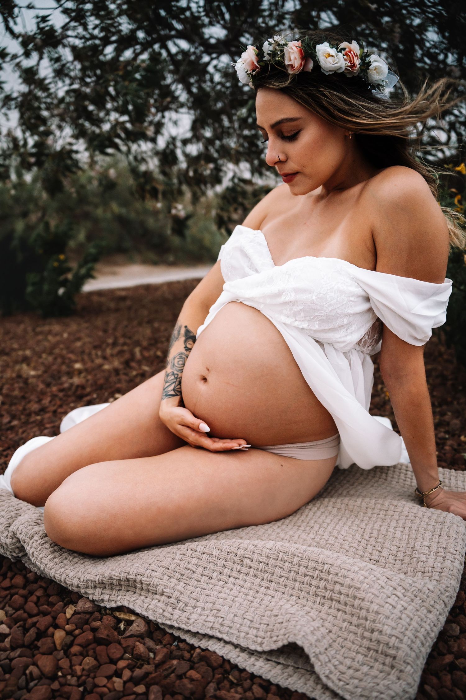 Pregnant woman in white off-shoulder top and floral crown sits outdoors gently holding her bare baby bump.