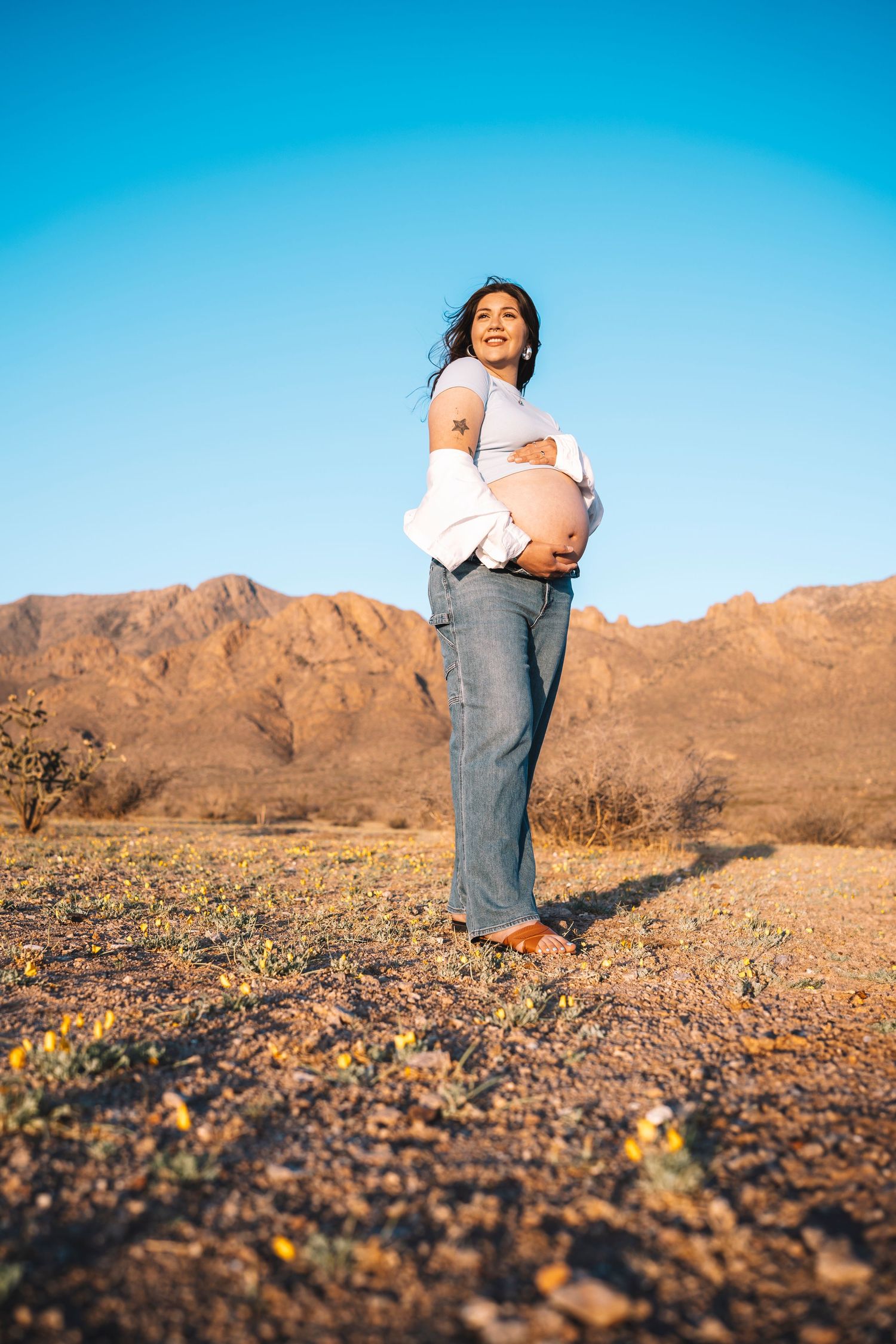Laughing pregnant woman holding bare baby bump barefoot in desert with mountain range behind her.