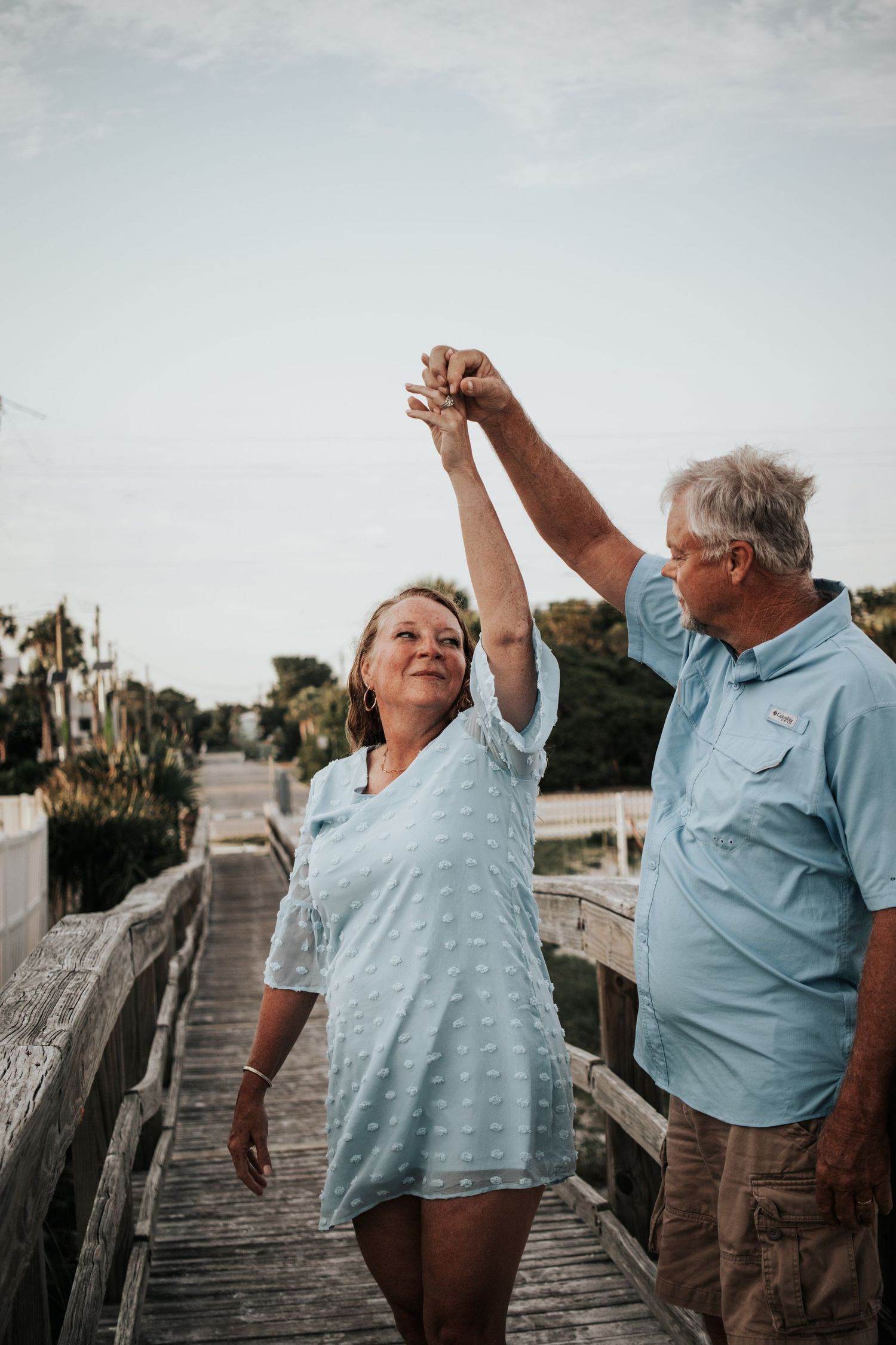 Older couple dancing on a boardwalk, man spinning woman by raised hands outdoors.