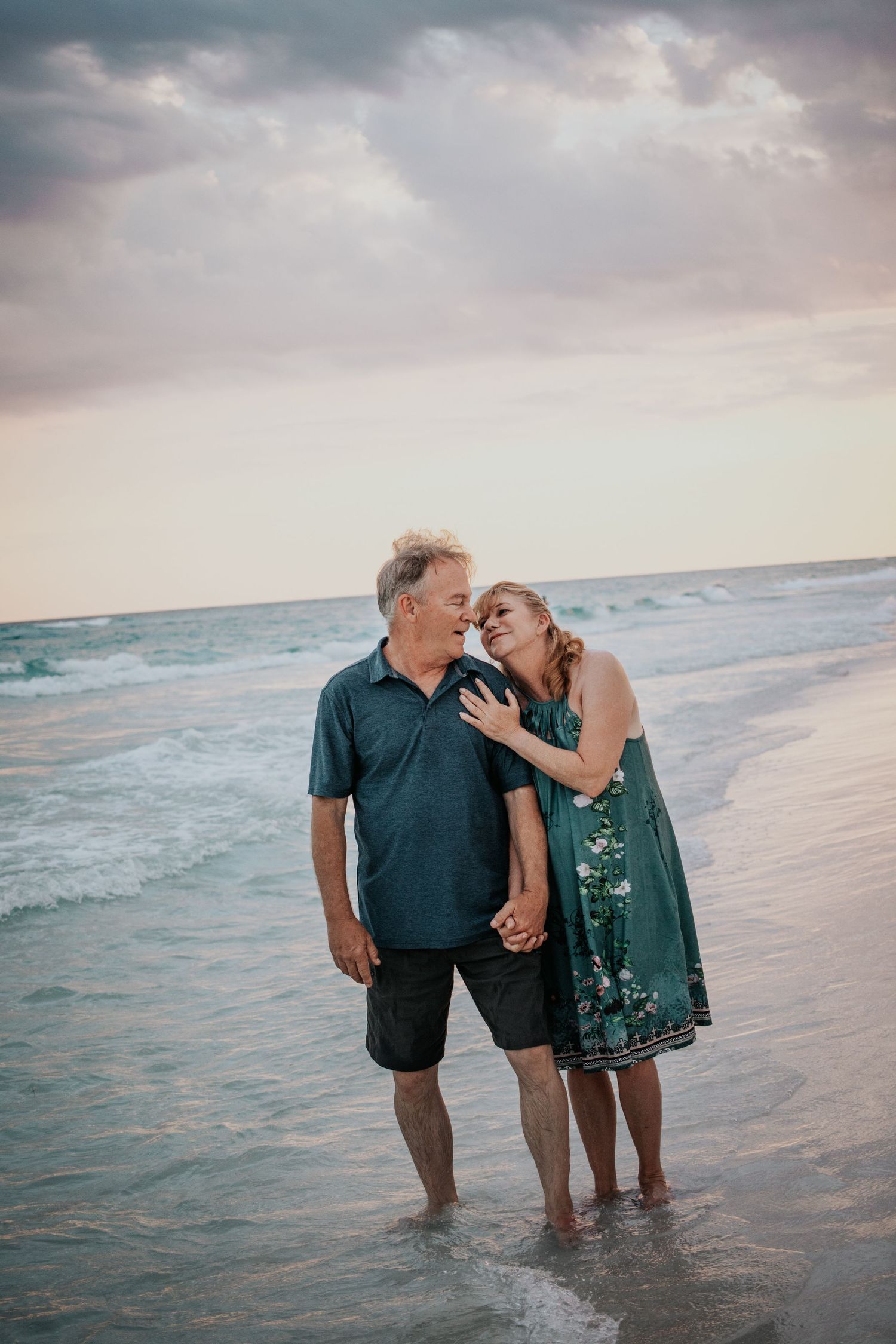 Couple laughing and leaning close together while standing in the ocean surf at sunset.