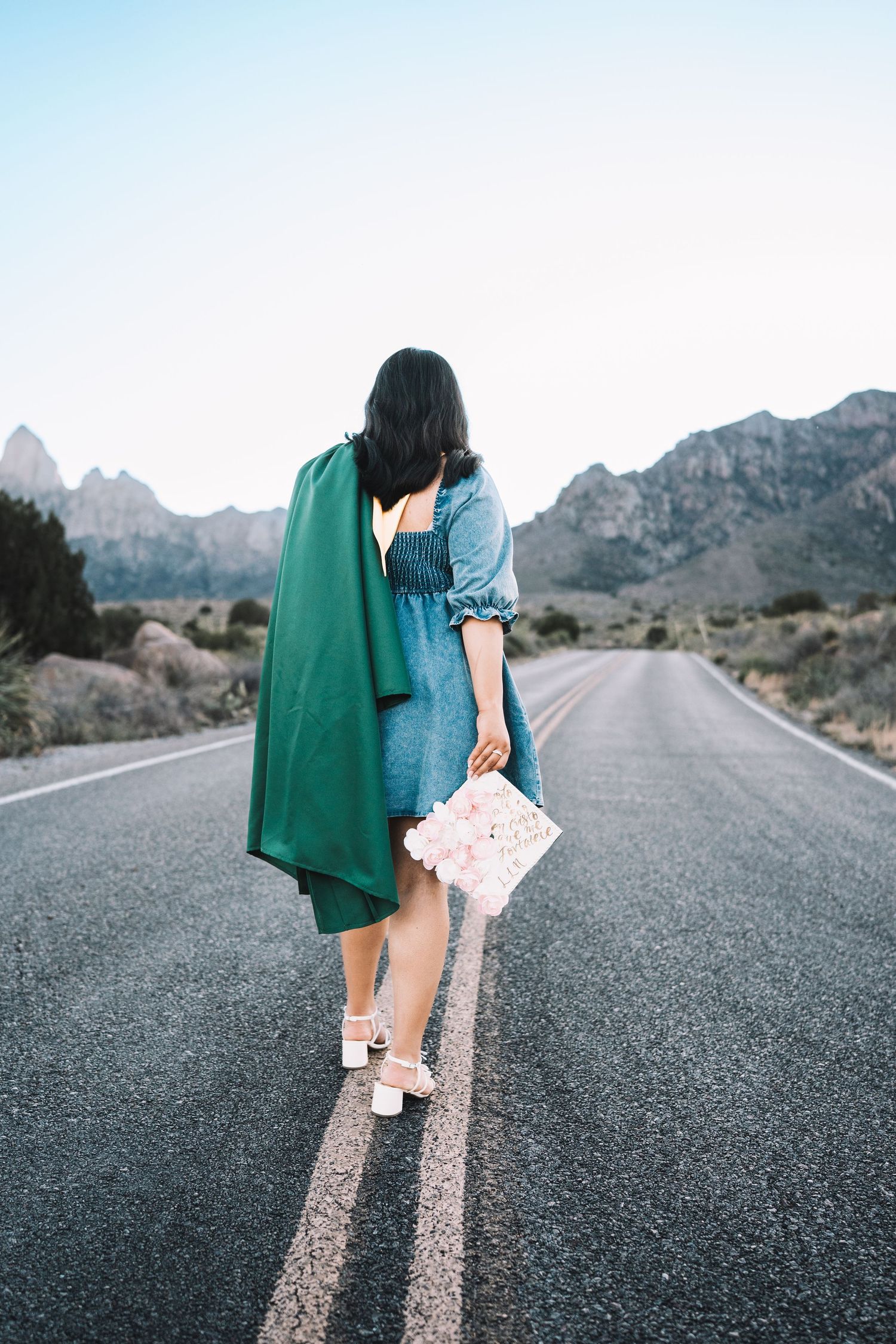 Graduate standing on a desert road holding a green gown and floral-decorated graduation cap with mountains behind.