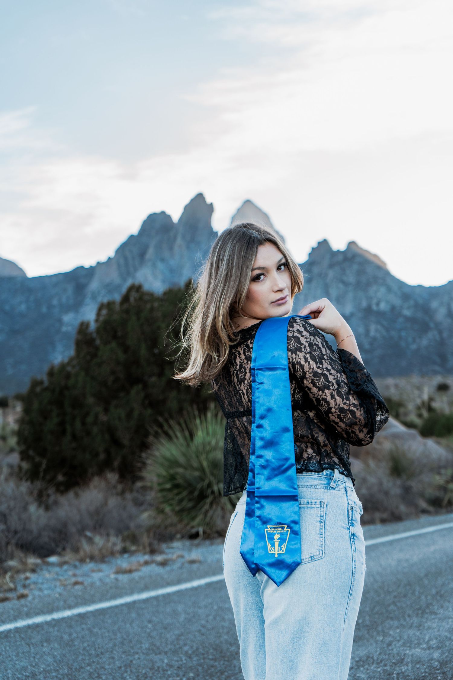 Young woman in black lace top displays blue graduation stole on desert mountain road.