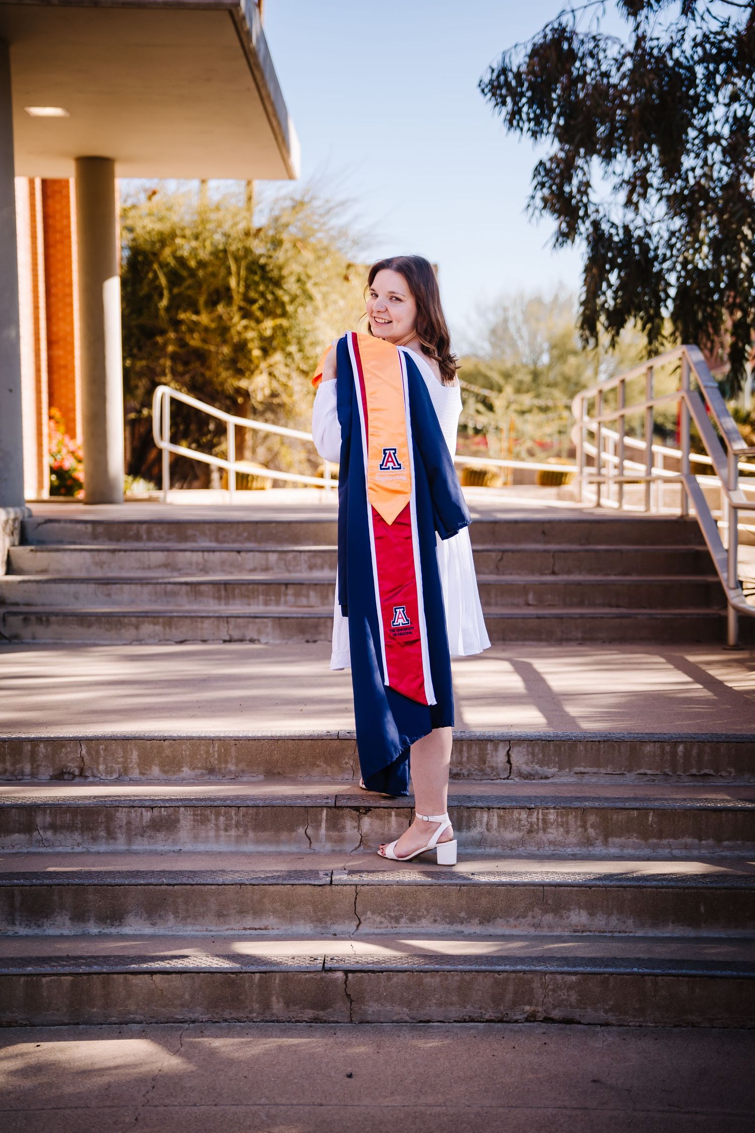 Female graduate in navy blue cap and gown with Arizona Wildcats stole on campus steps.