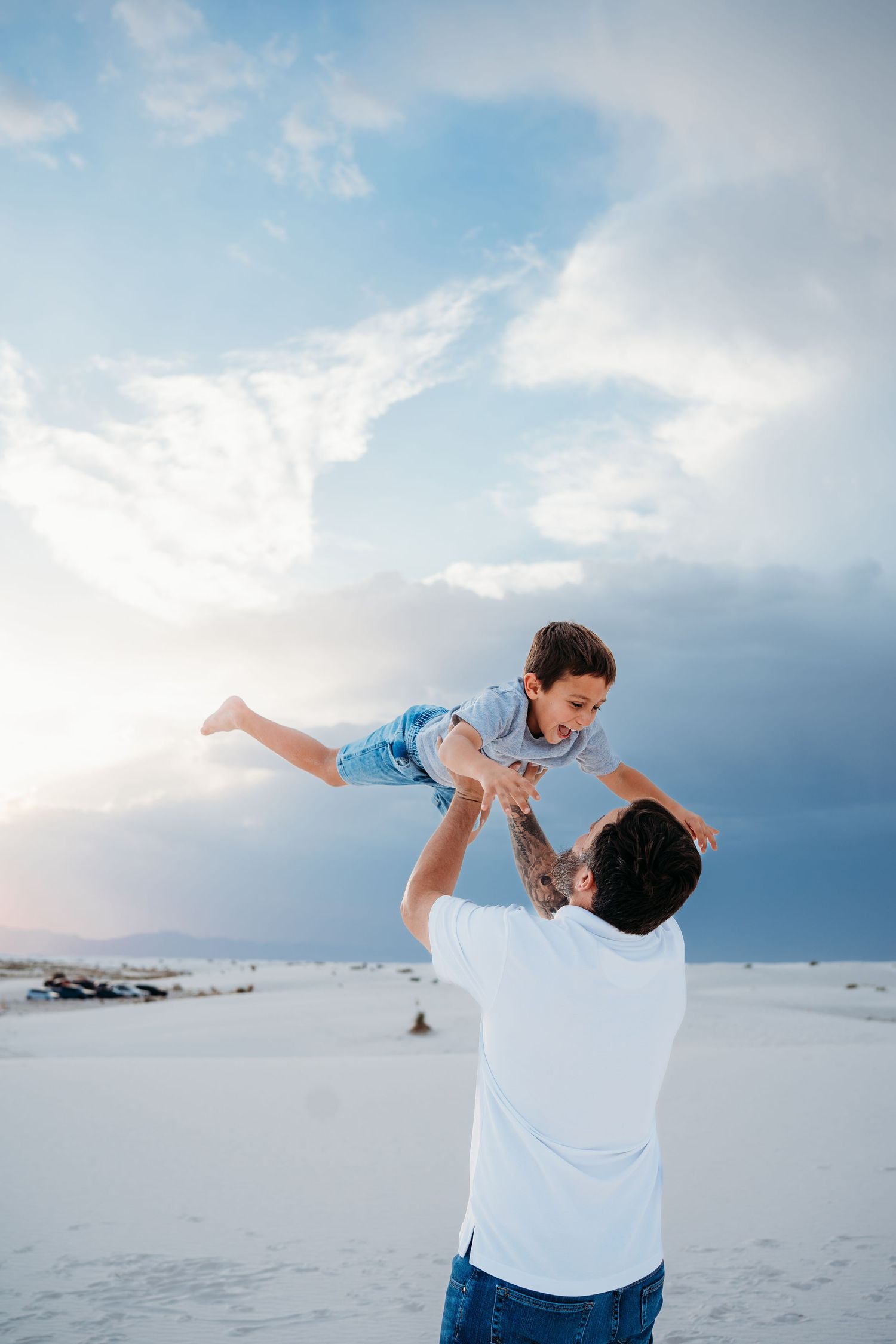A father tosses his laughing young son into the air on white sand dunes at sunset.