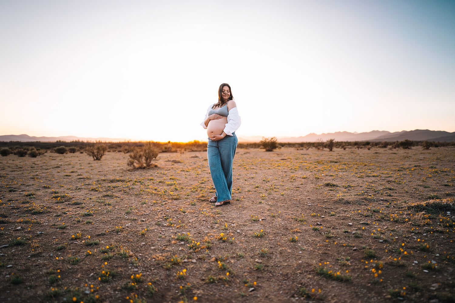 Pregnant woman standing in vast desert landscape holding belly at sunset with mountains.