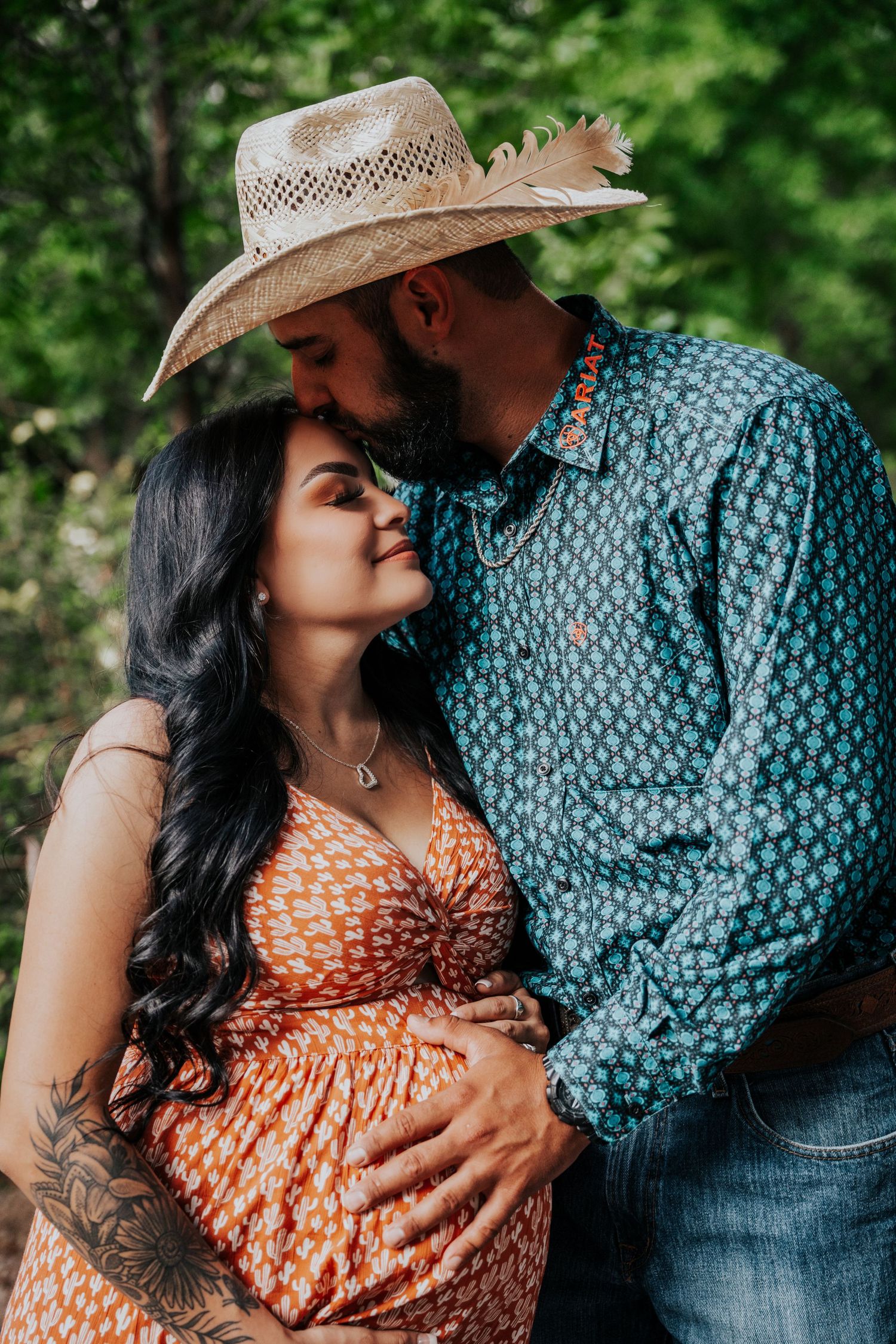 Couple sharing tender moment, pregnant woman in orange dress and tattooed arm, man in cowboy hat kisses her forehead.
