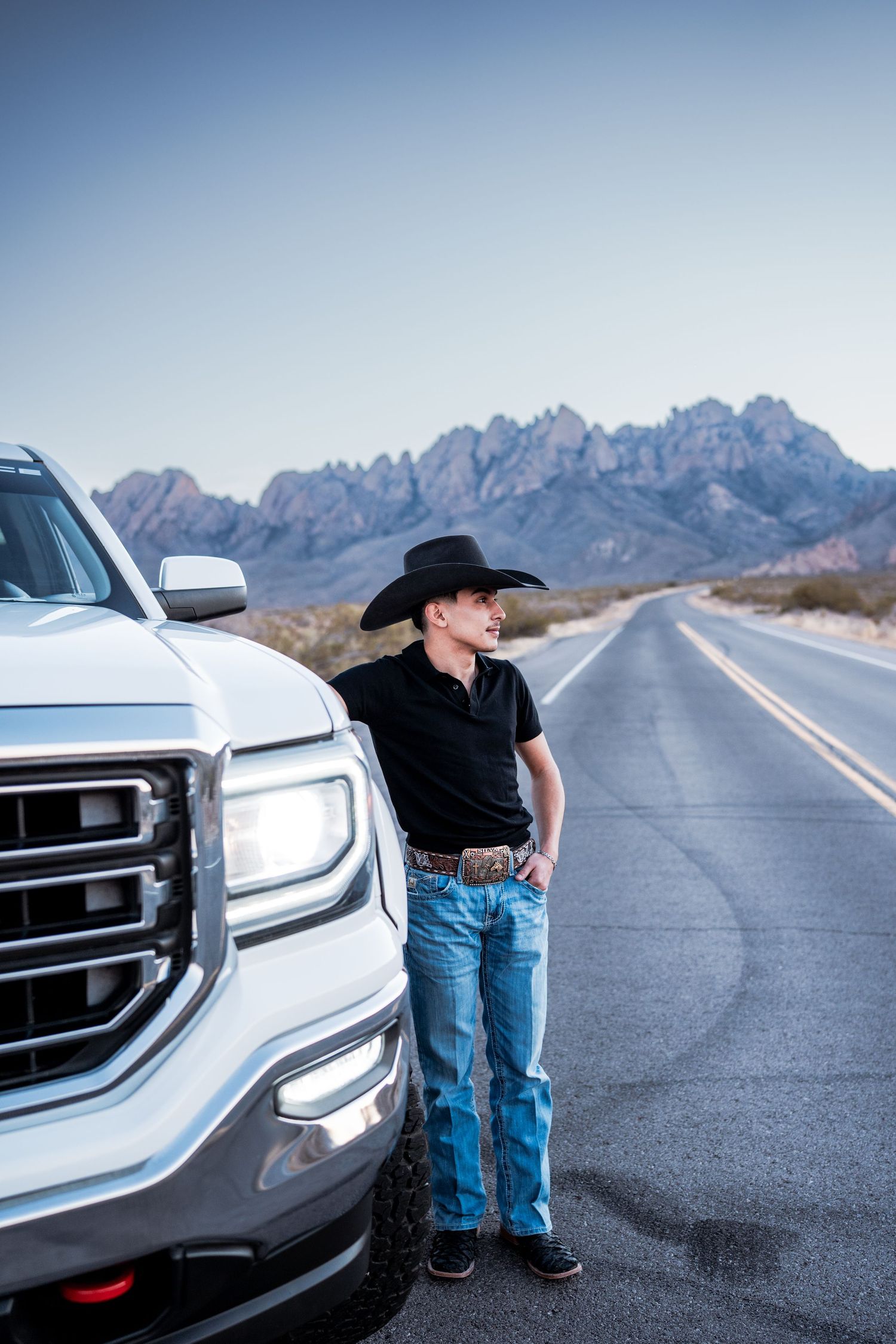 Young man in black cowboy hat stands beside white pickup truck on rural desert road with mountains.