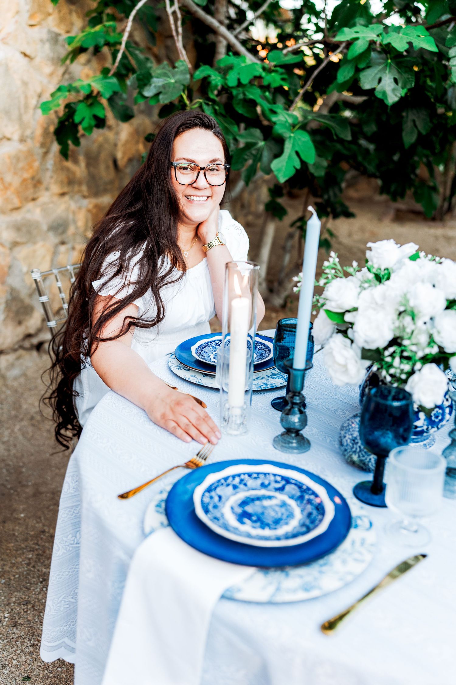 Elegant woman in white dress smiling at blue and white outdoor tablescape with white floral centerpiece and candles.