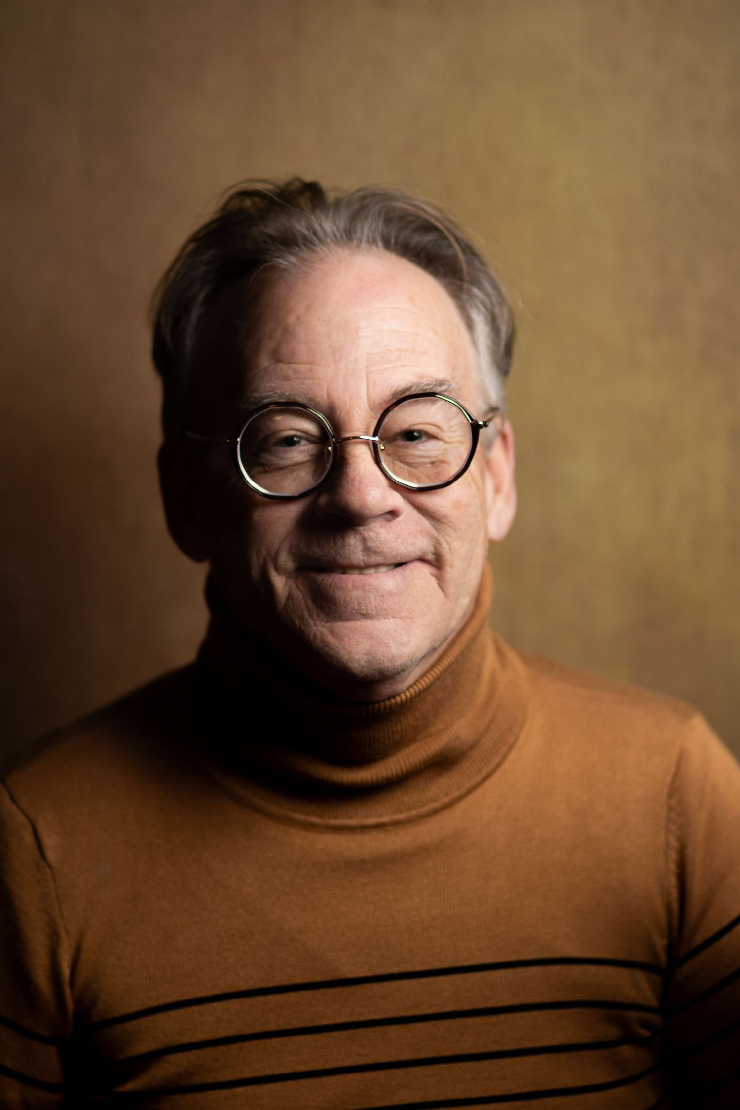 Smiling older man wearing round glasses and brown striped turtleneck sweater.