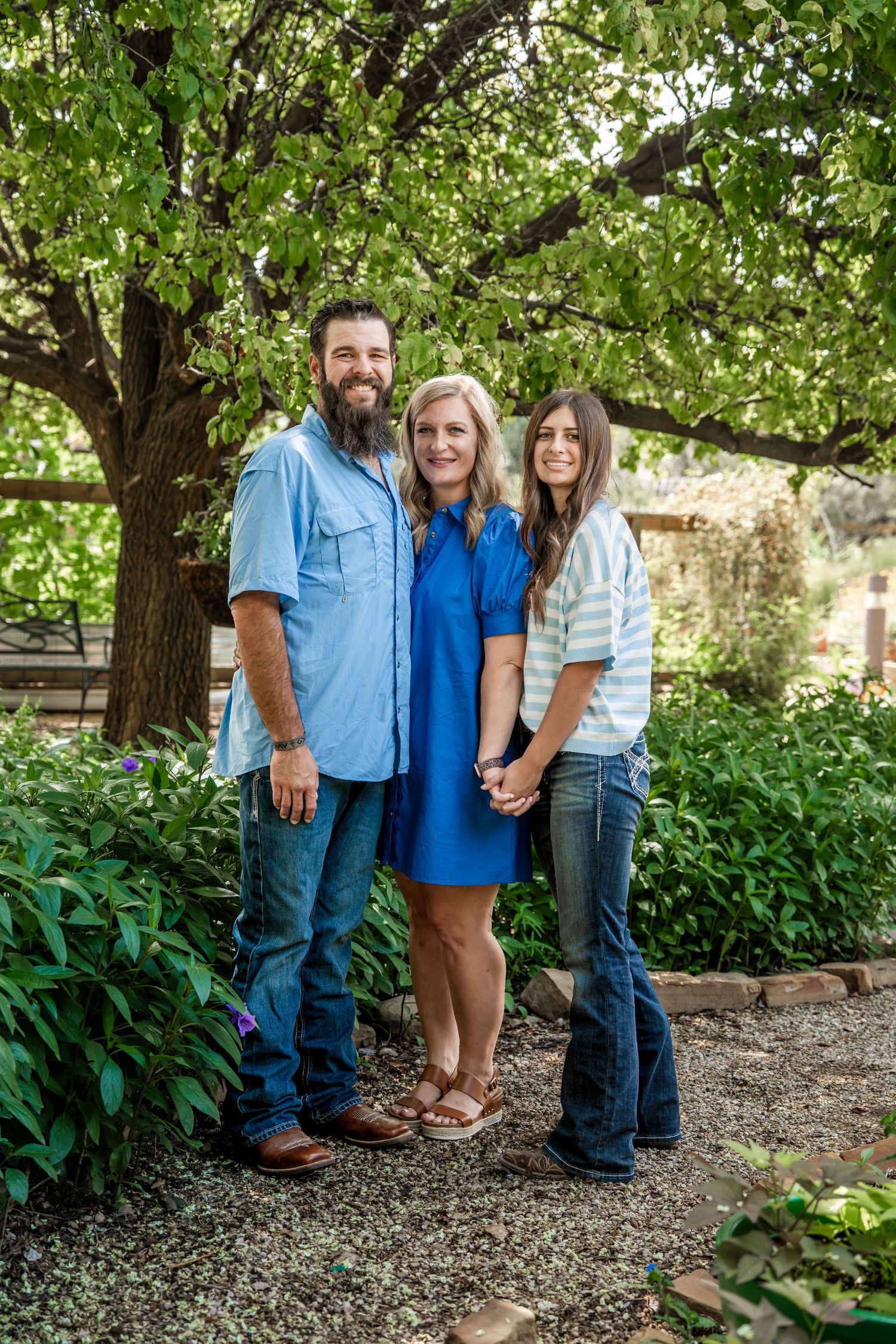 Man with beard, blonde woman in blue dress, and teen girl posing in garden.