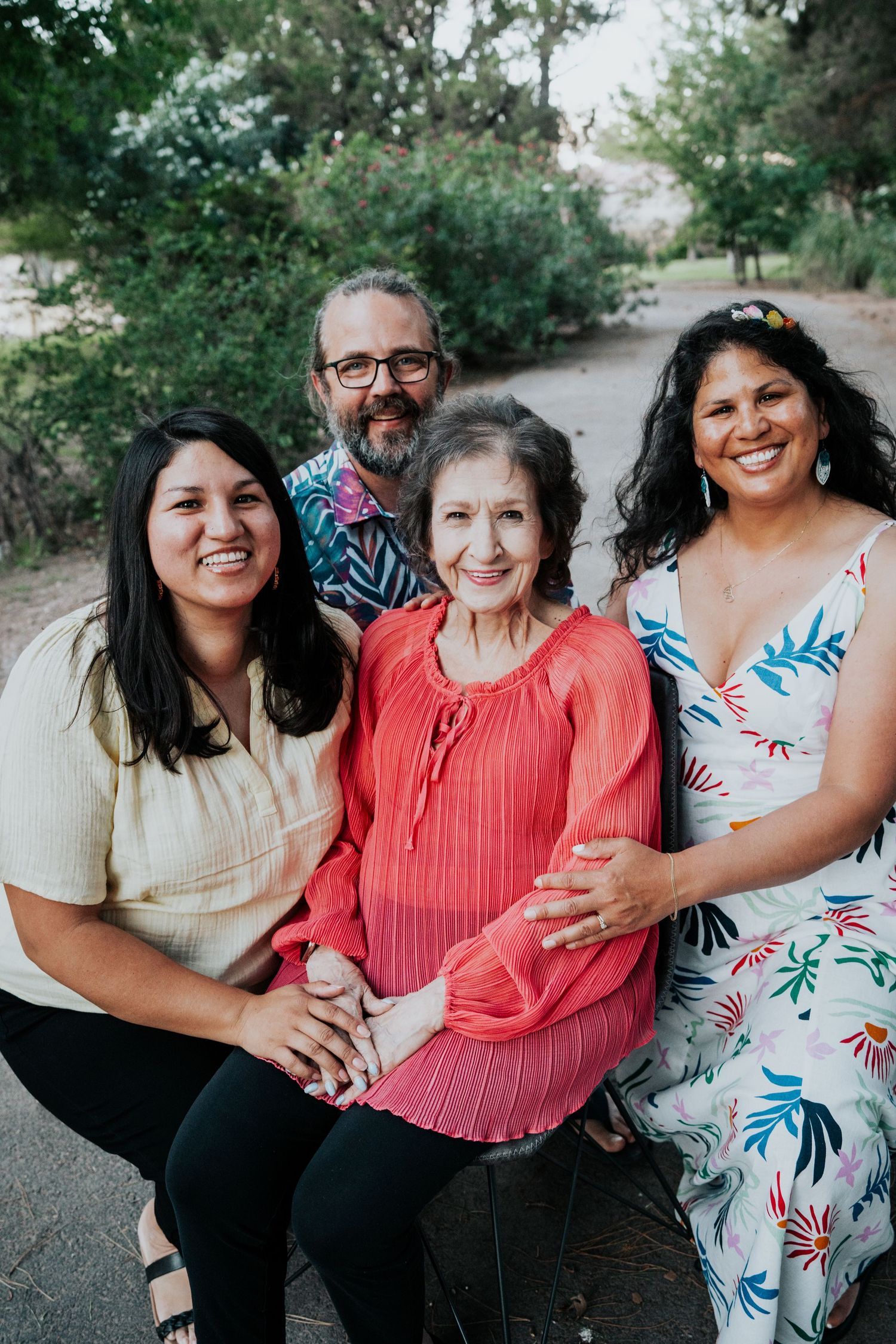 Family of four smiles together outdoors, with an elderly woman seated in the center of the group.
